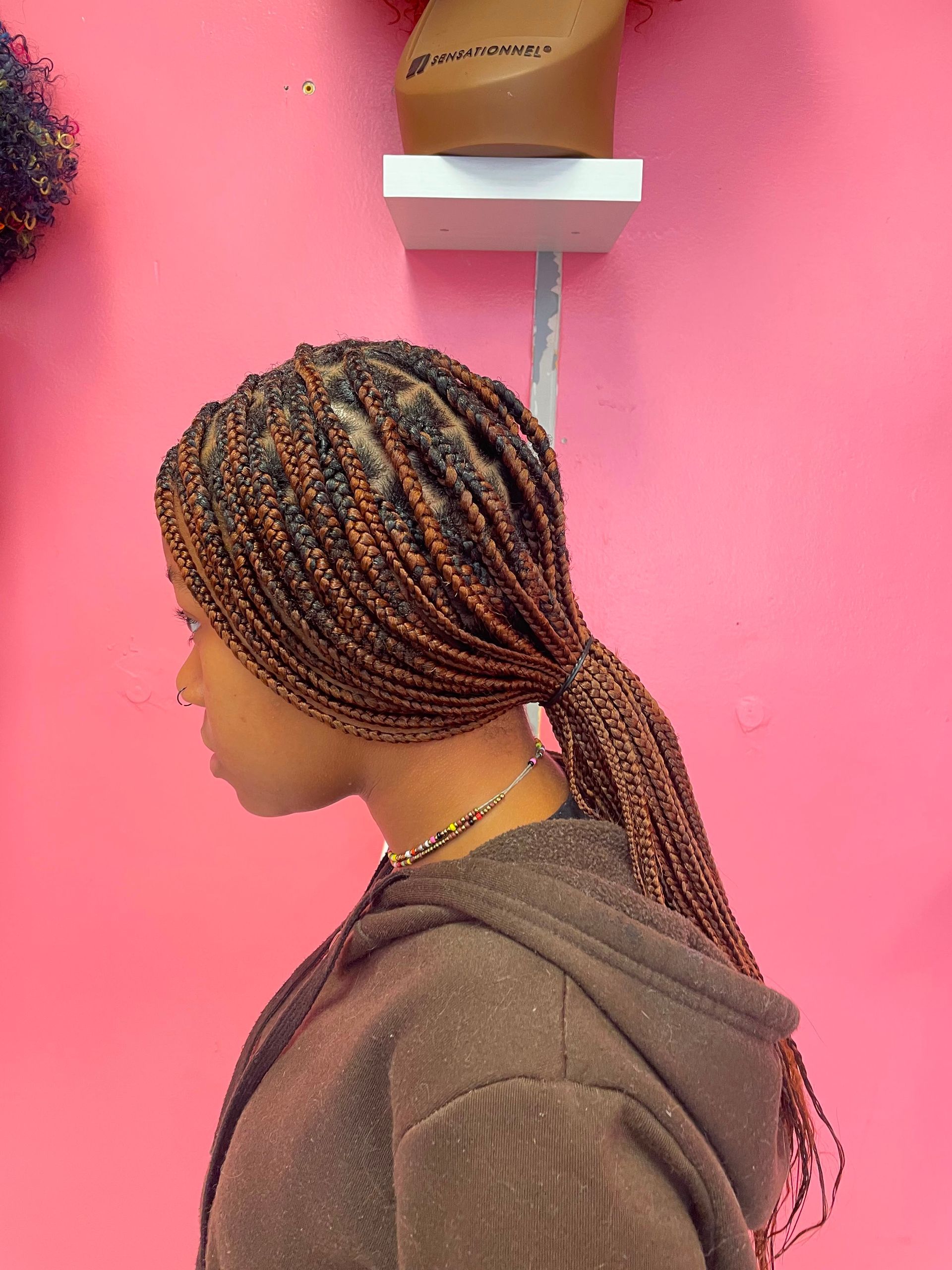 Side profile of a person with braided hair against a bright pink wall under a wall-mounted dryer