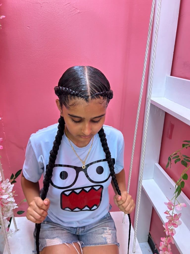 Girl with braided hair sitting on a swing in a pink room, wearing a cartoon face T-shirt and denim shorts
