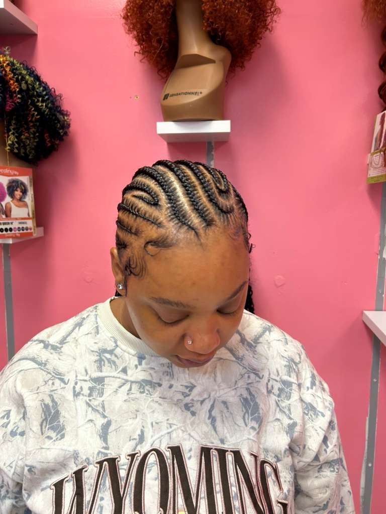 Person with braided hair looking down in a pink salon, wearing a white shirt with “WOMING.”