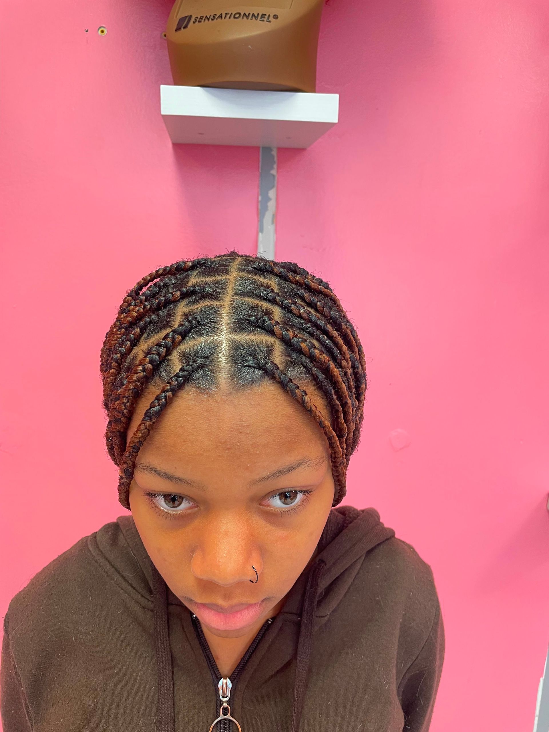 Person with braided hair looking down against a pink wall, under a shelf with a brown box.