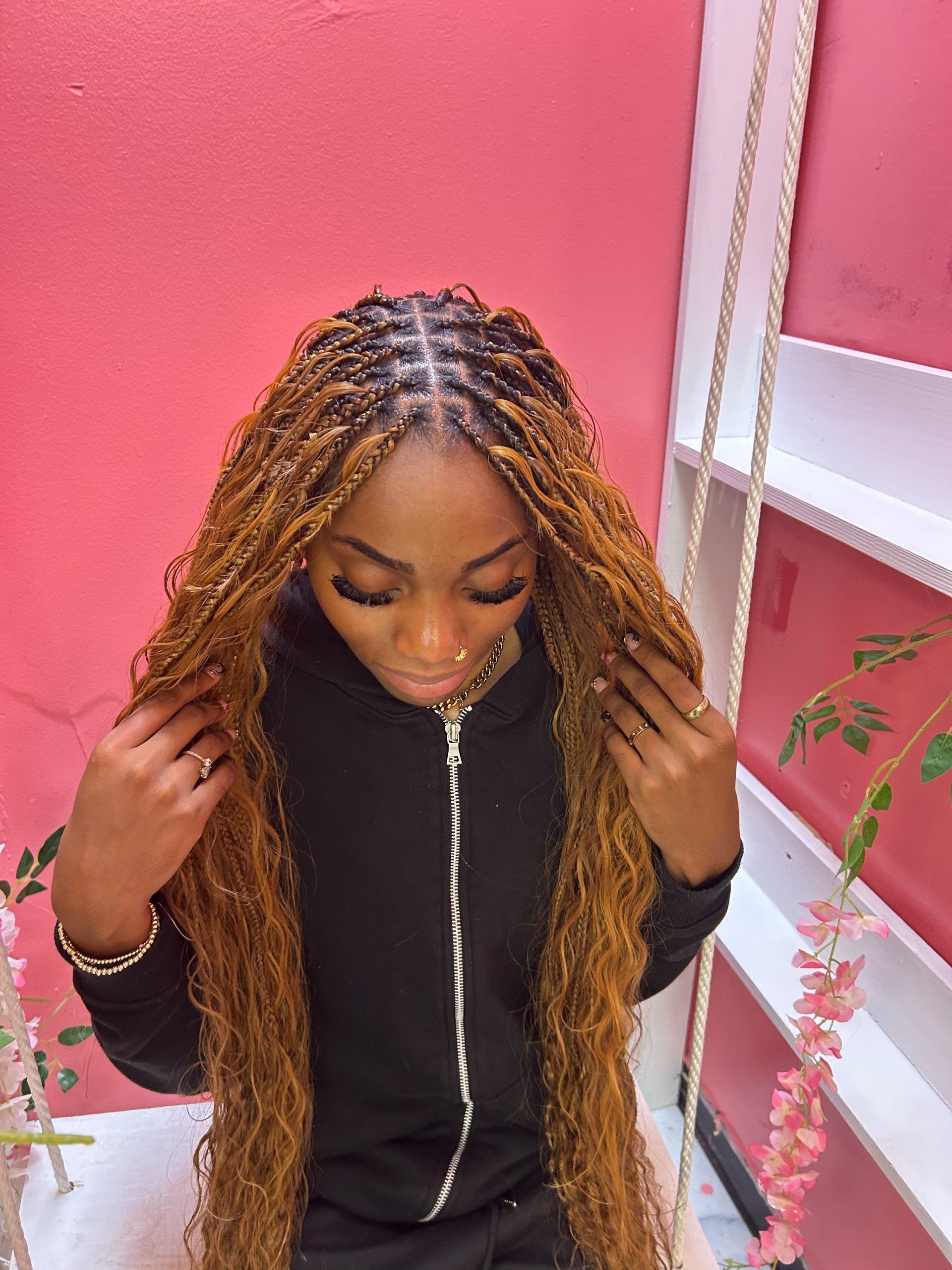 Person with long braided hair looking down in a black top against a pink wall and white shelf.