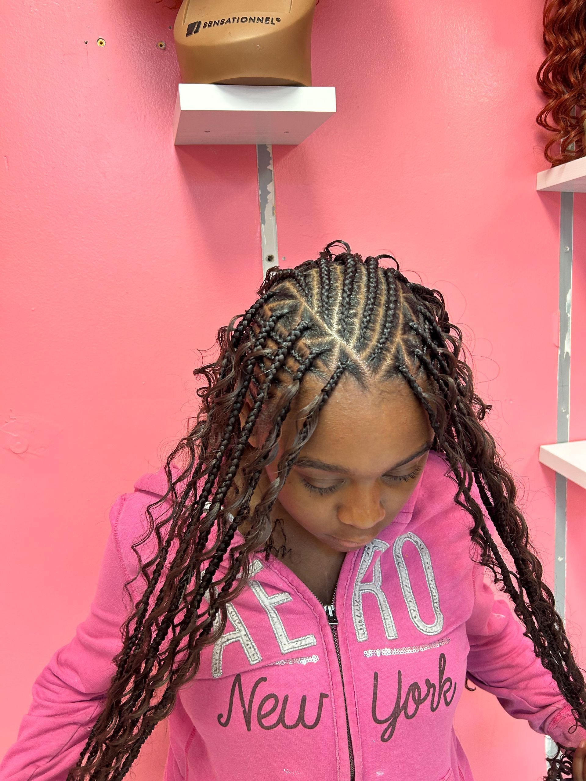Girl with braided hair looking down, wearing a pink New York hoodie against a pink wall.