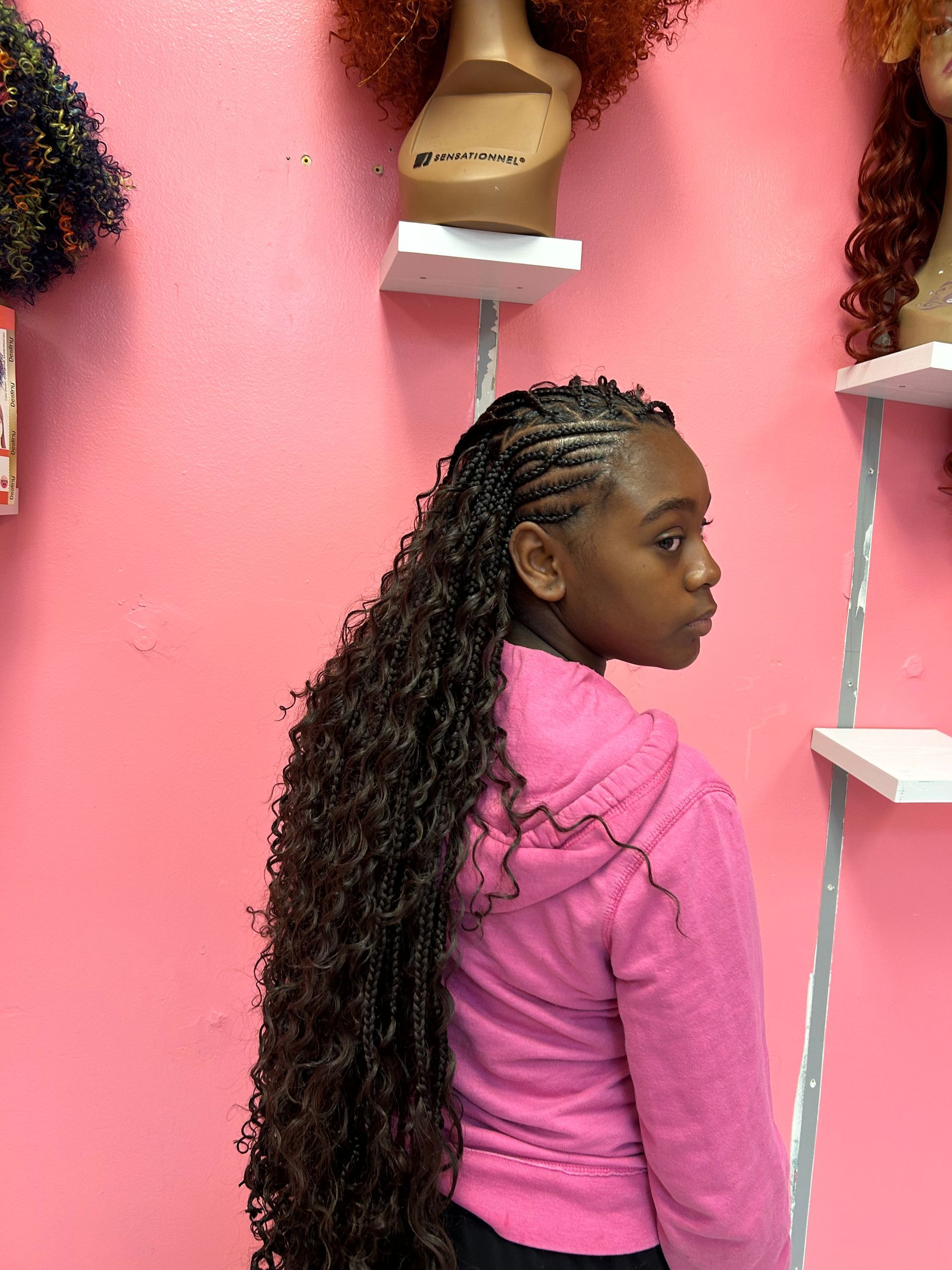 Woman with long braided hair in a pink top, standing in profile against a pink wall with shelves.