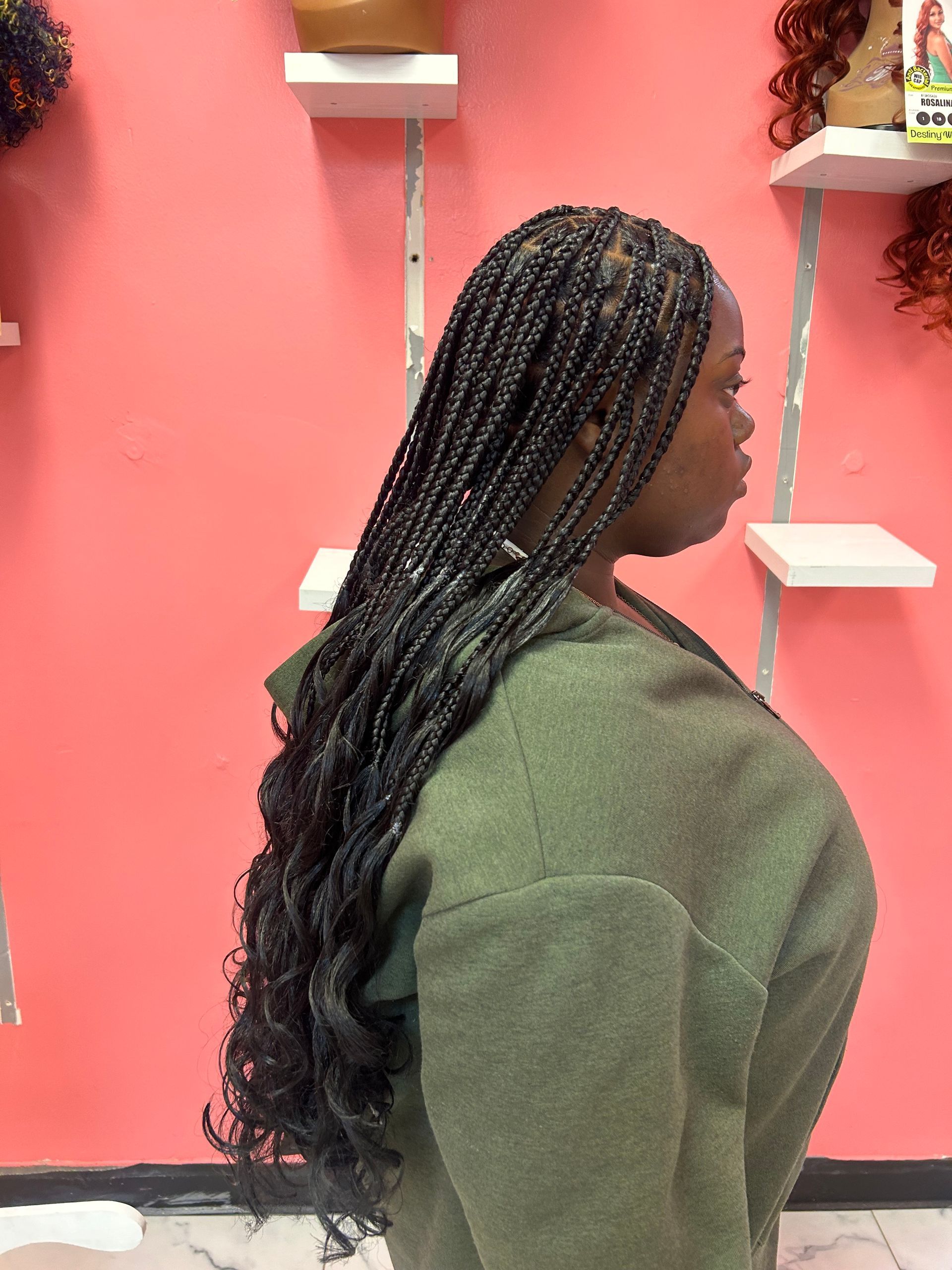 Side-profile person with long braided hair wearing a green sweater against a pink wall with shelves.