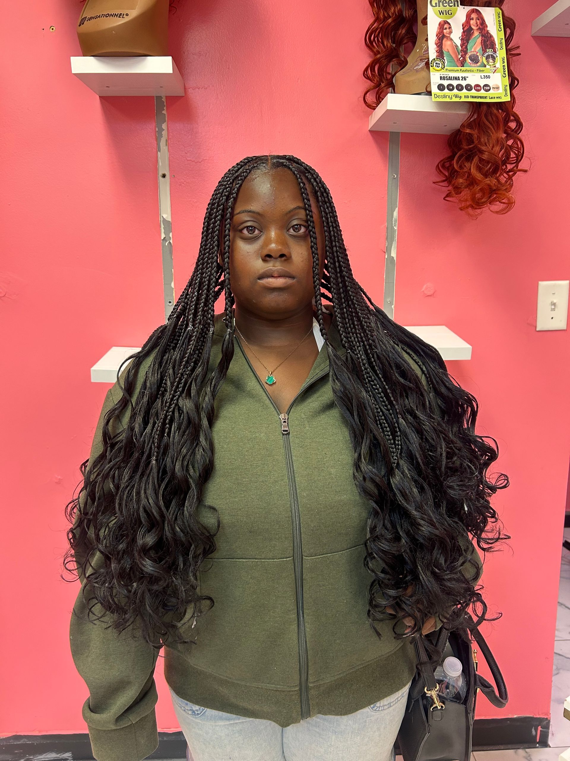 Person with long braided hair stands against a pink wall, wearing a green zip-up sweater.