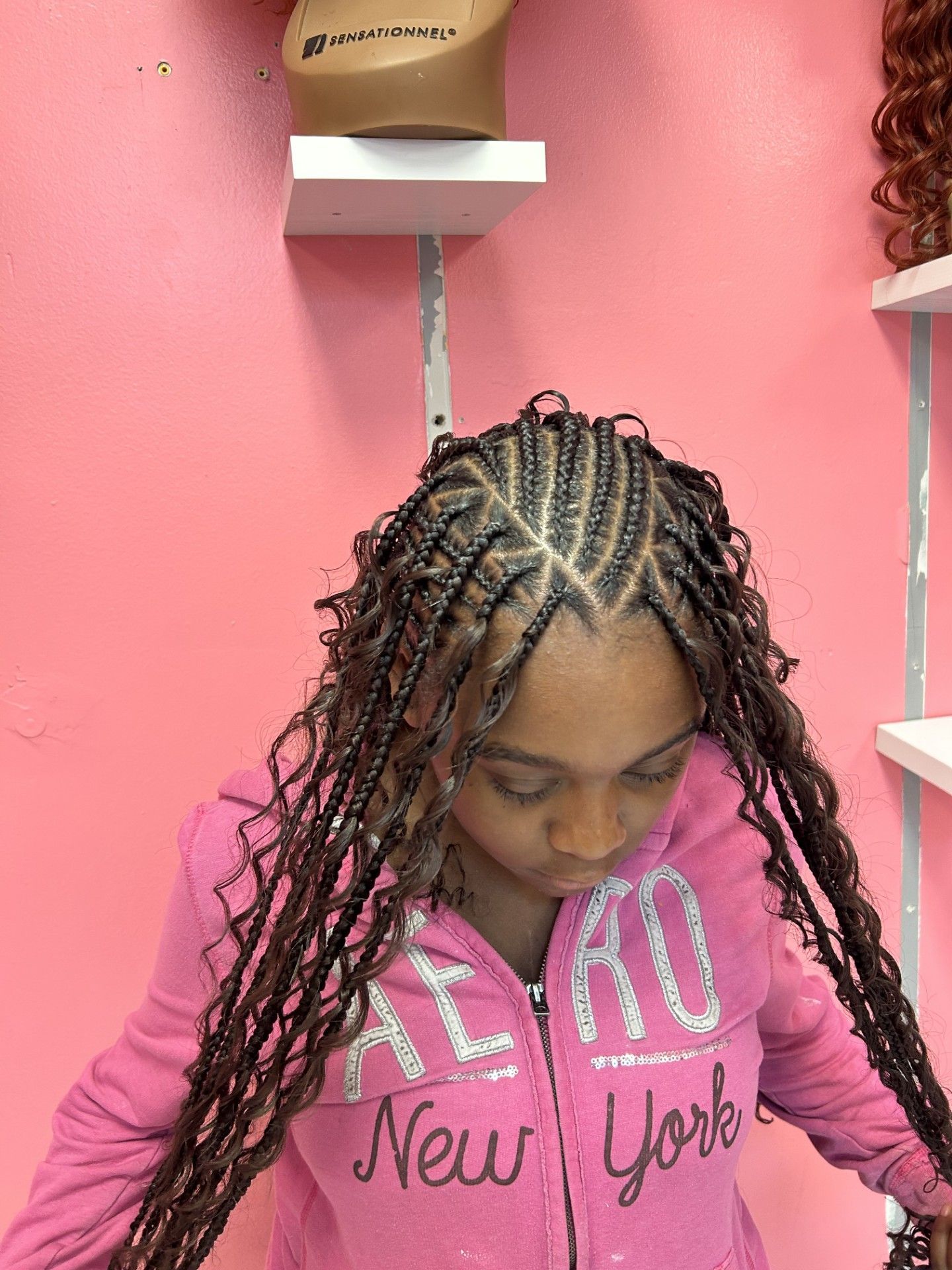 Person with braided hair looking down in a pink room, wearing a pink “New York” hoodie.
