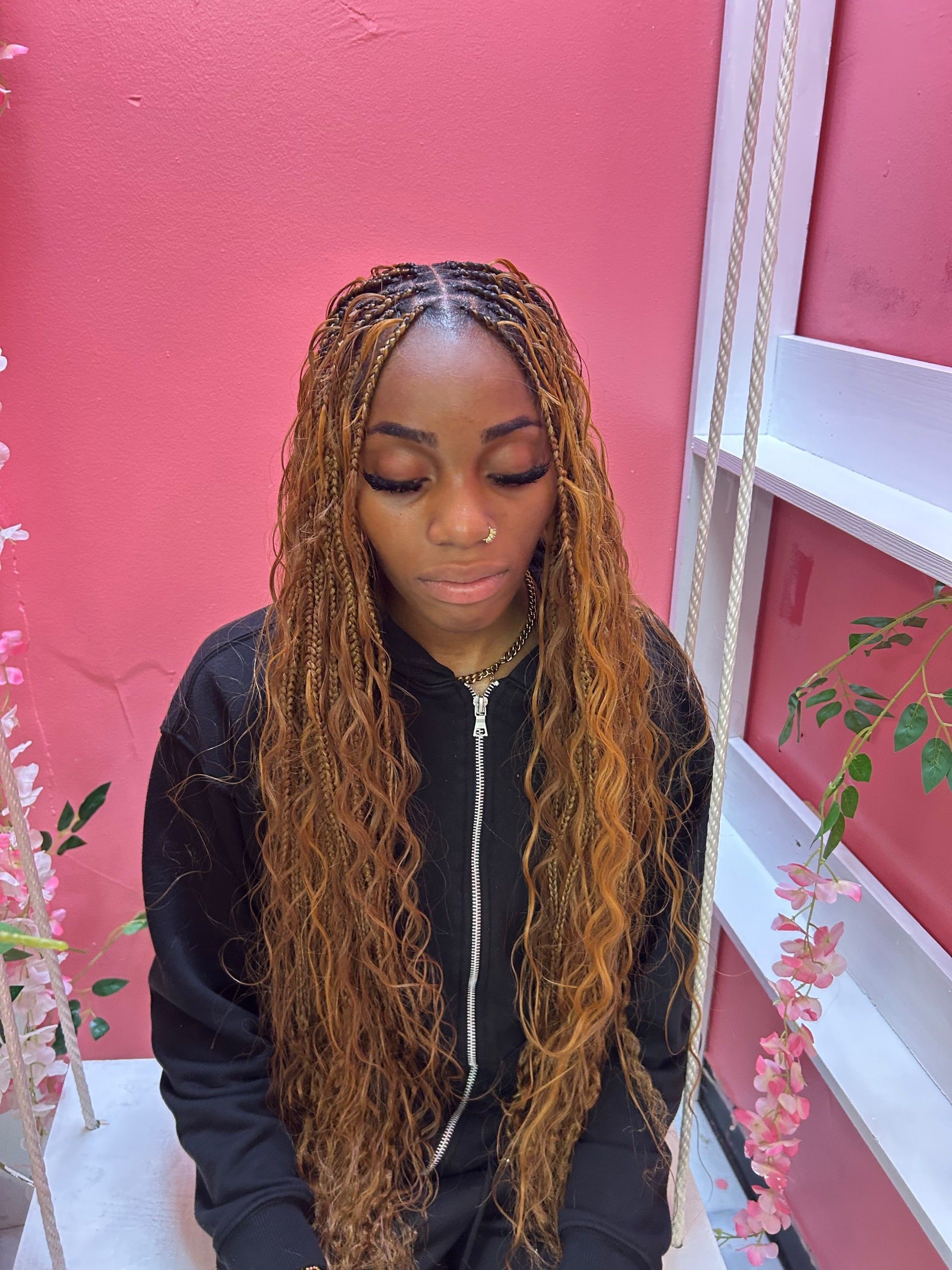 Woman with long braids in a black hoodie, standing against a pink wall and looking down.