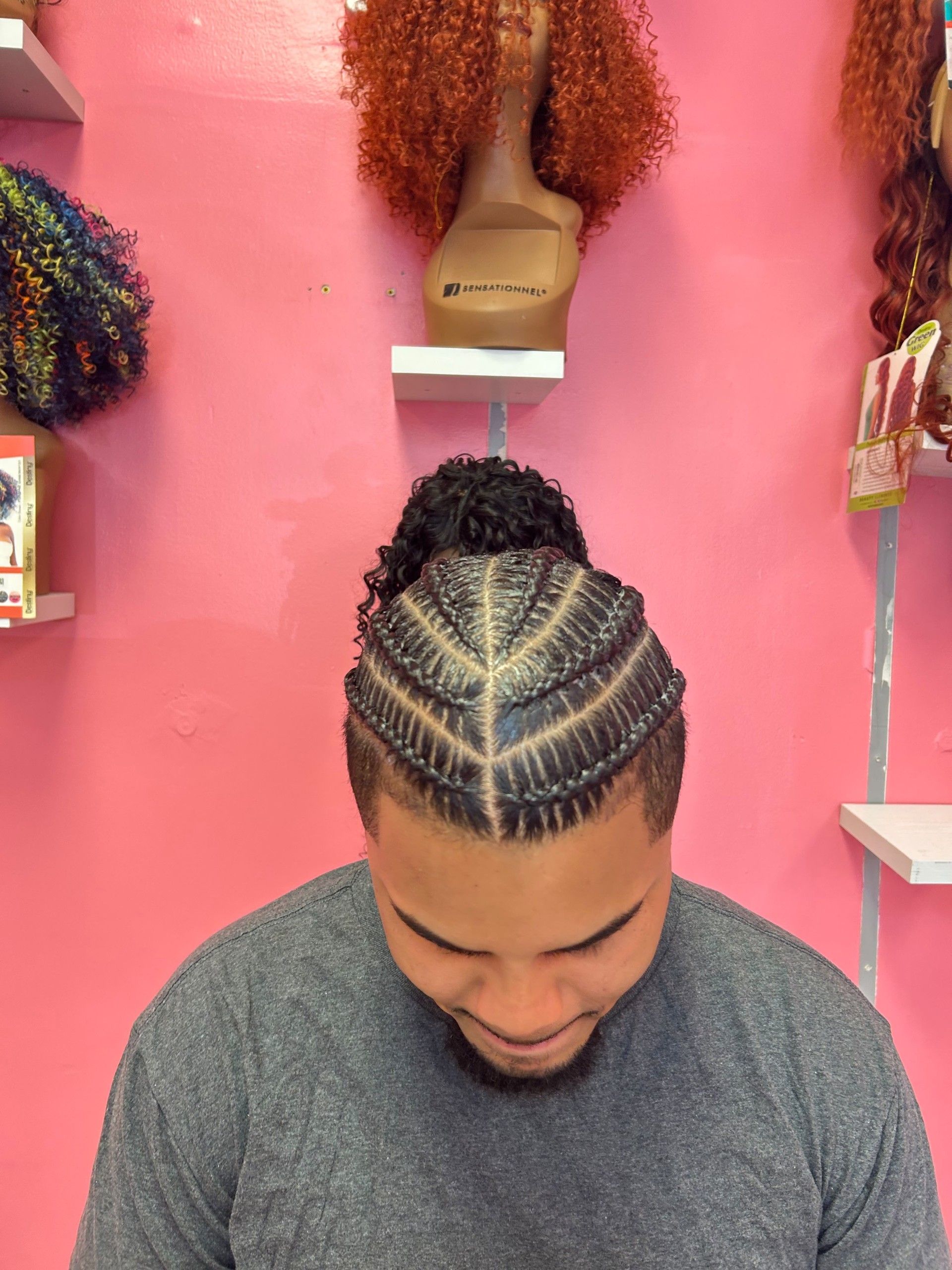 Person with braided hair looking down in a pink salon with wig displays on the wall