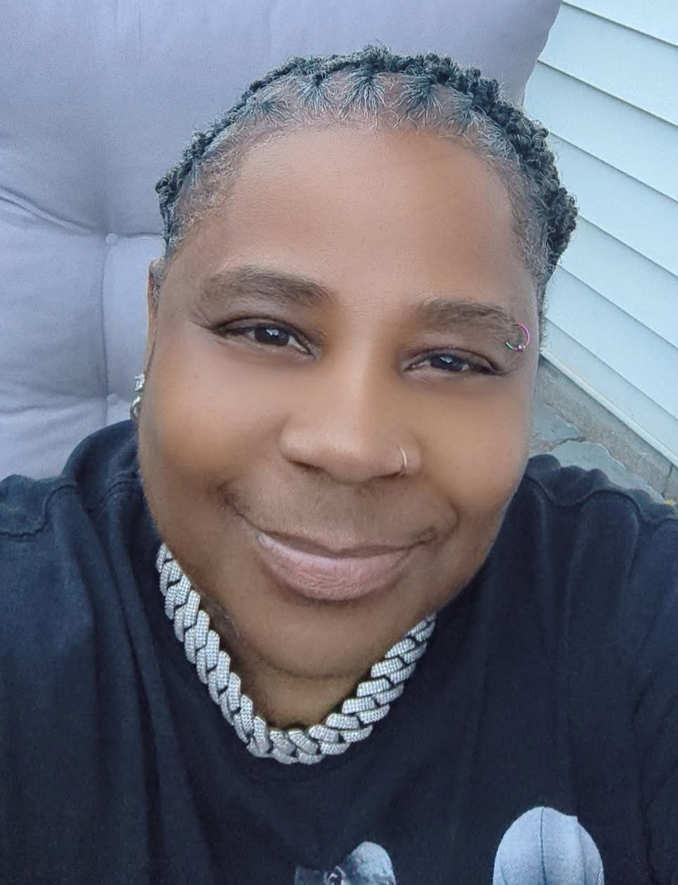 Person smiling outdoors in a black shirt and silver chain necklace, seated beside a light gray cushion.