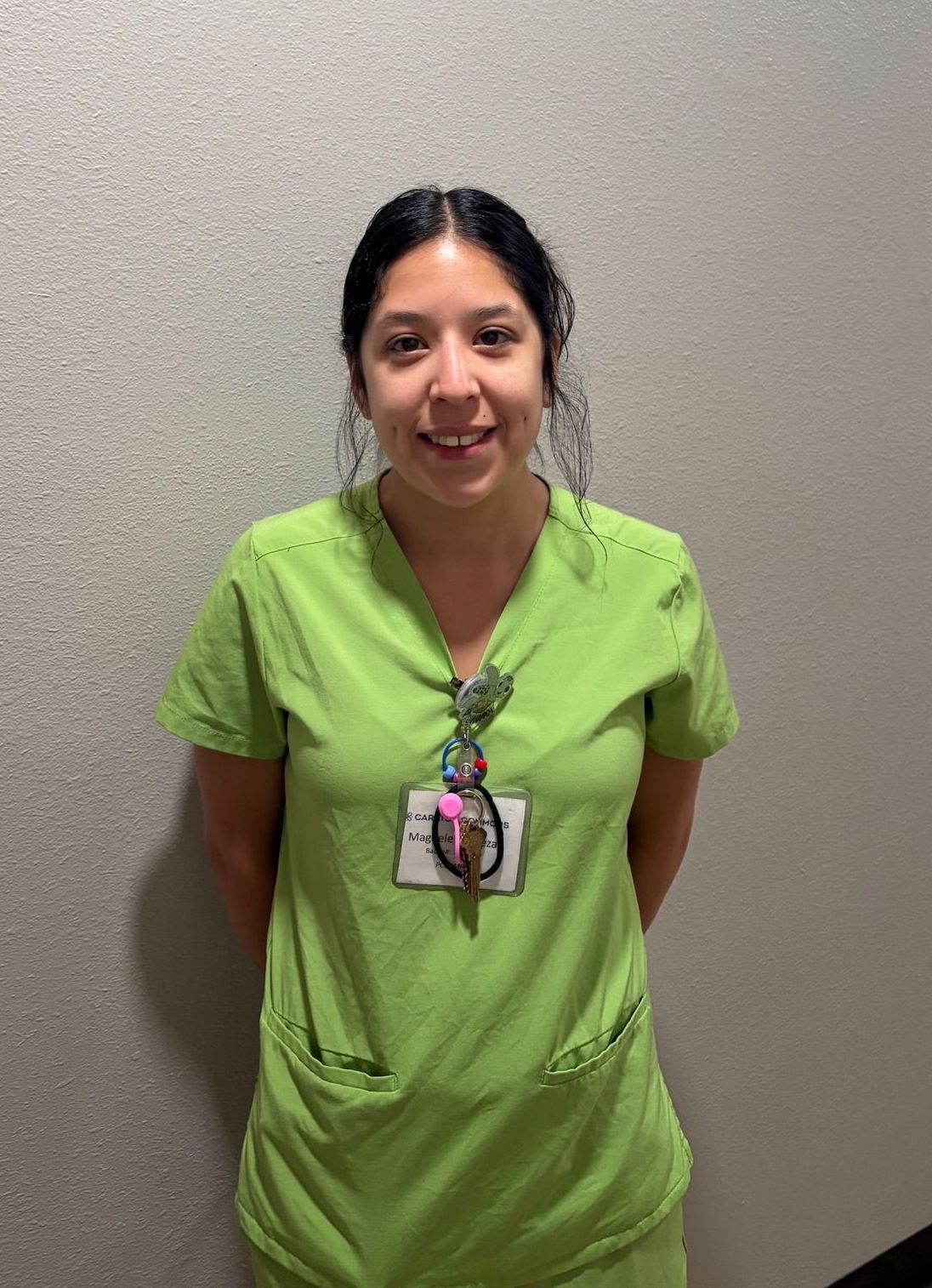 Person in green scrubs with a lanyard, standing against a gray wall and smiling.