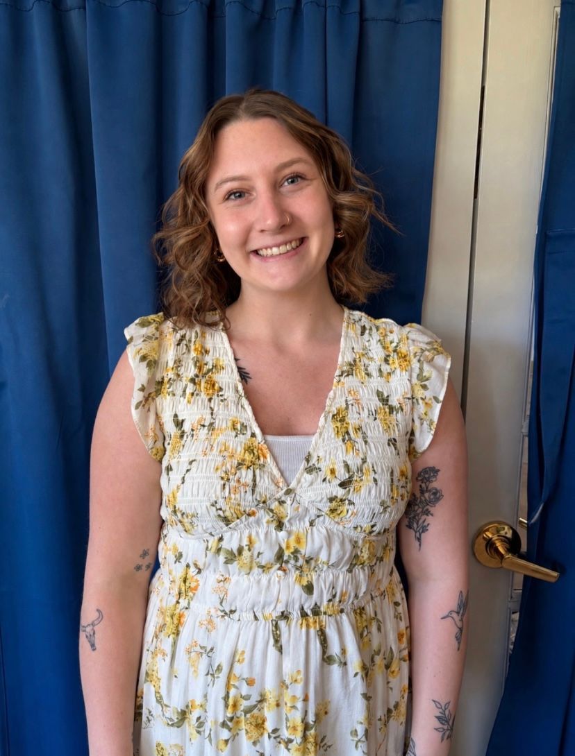 Smiling person in navy scrubs with a badge, standing against a dotted beige wall.