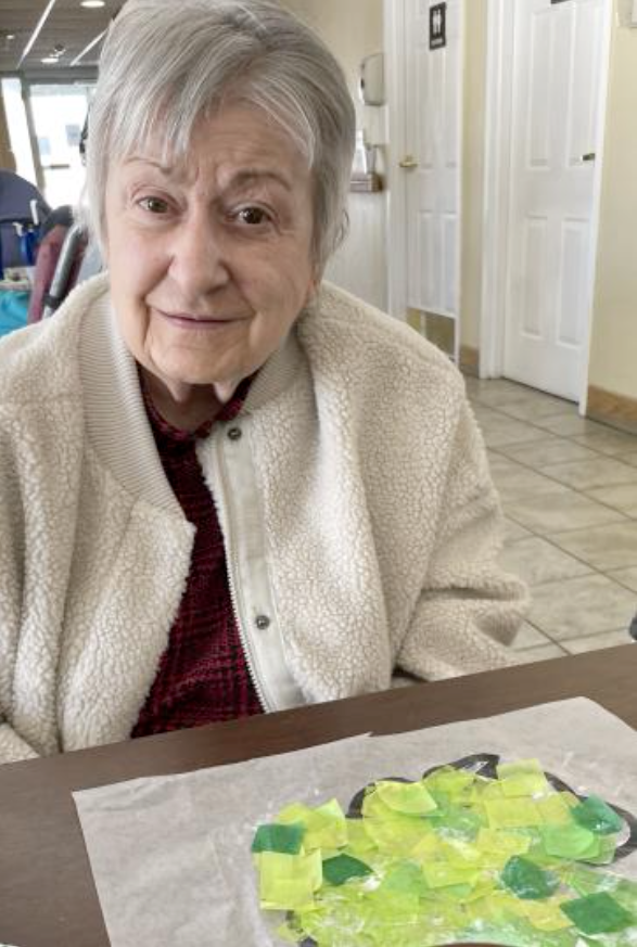 An elderly woman is sitting at a table with a painting on it.