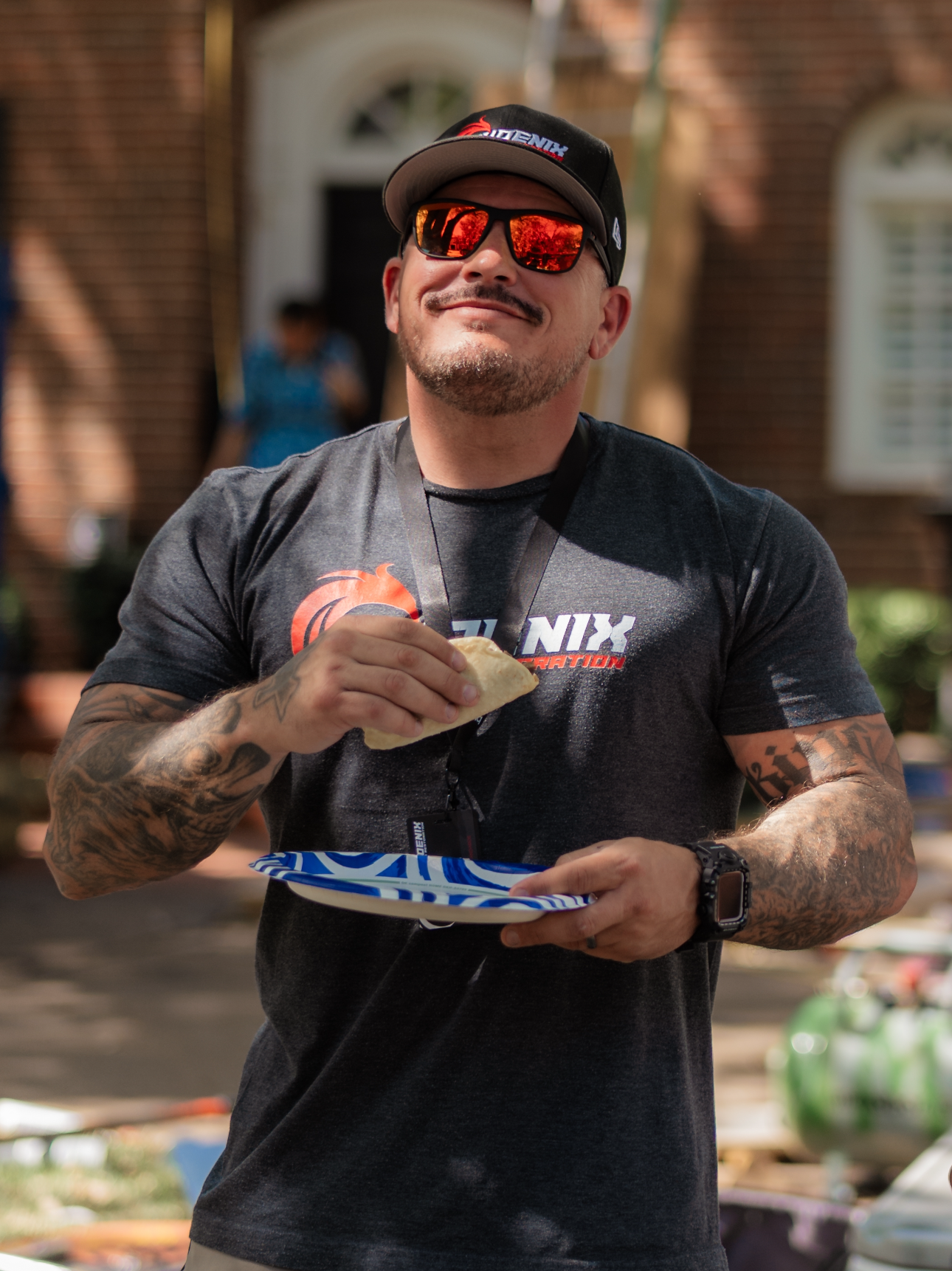 Man in cap and sunglasses eating a taco outside a brick building.