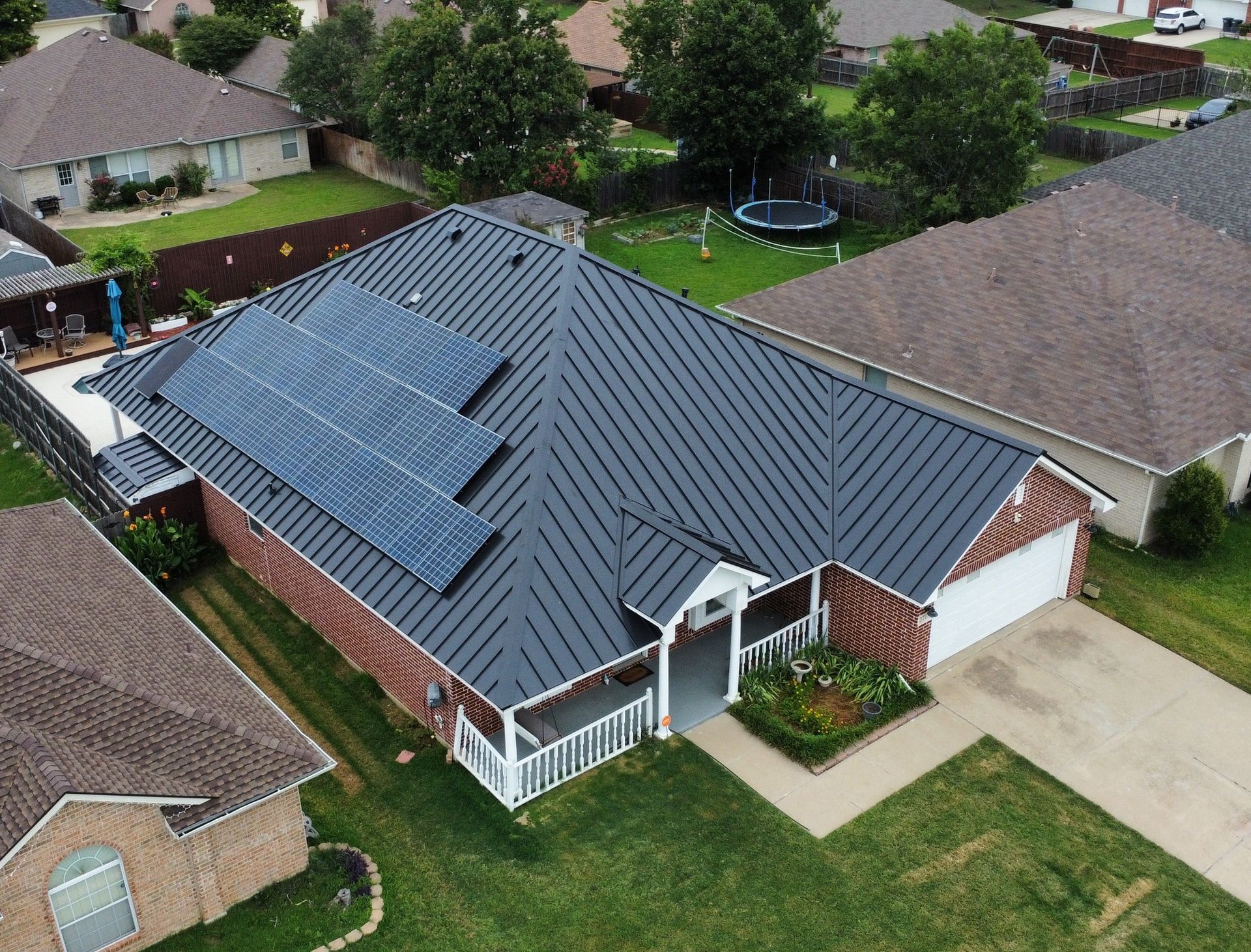 Aerial view of a house with solar panels on the dark metal roof, surrounded by lawns and other homes.