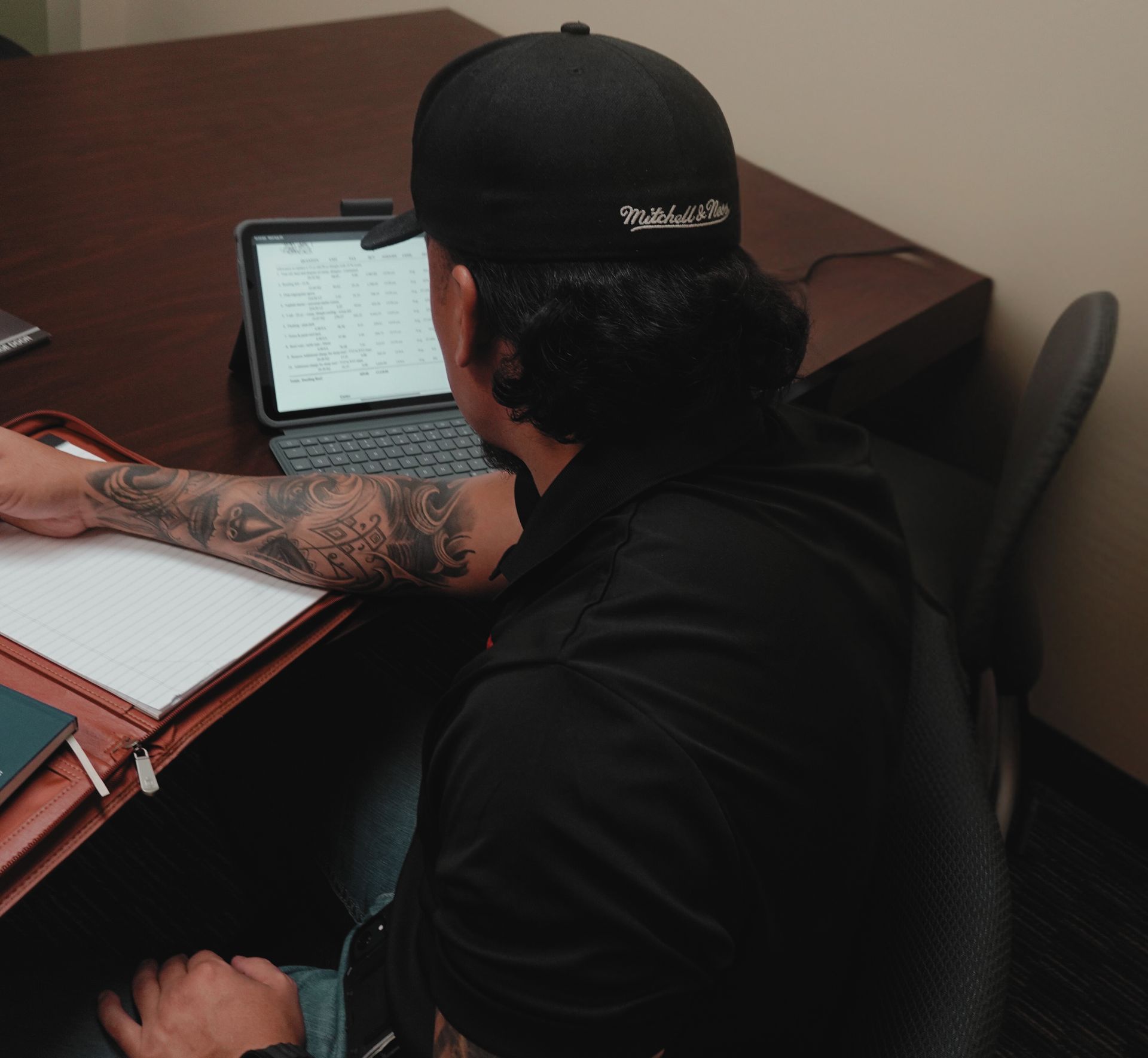 Man in black cap and shirt with arm tattoos working at a desk with tablet and notebook.