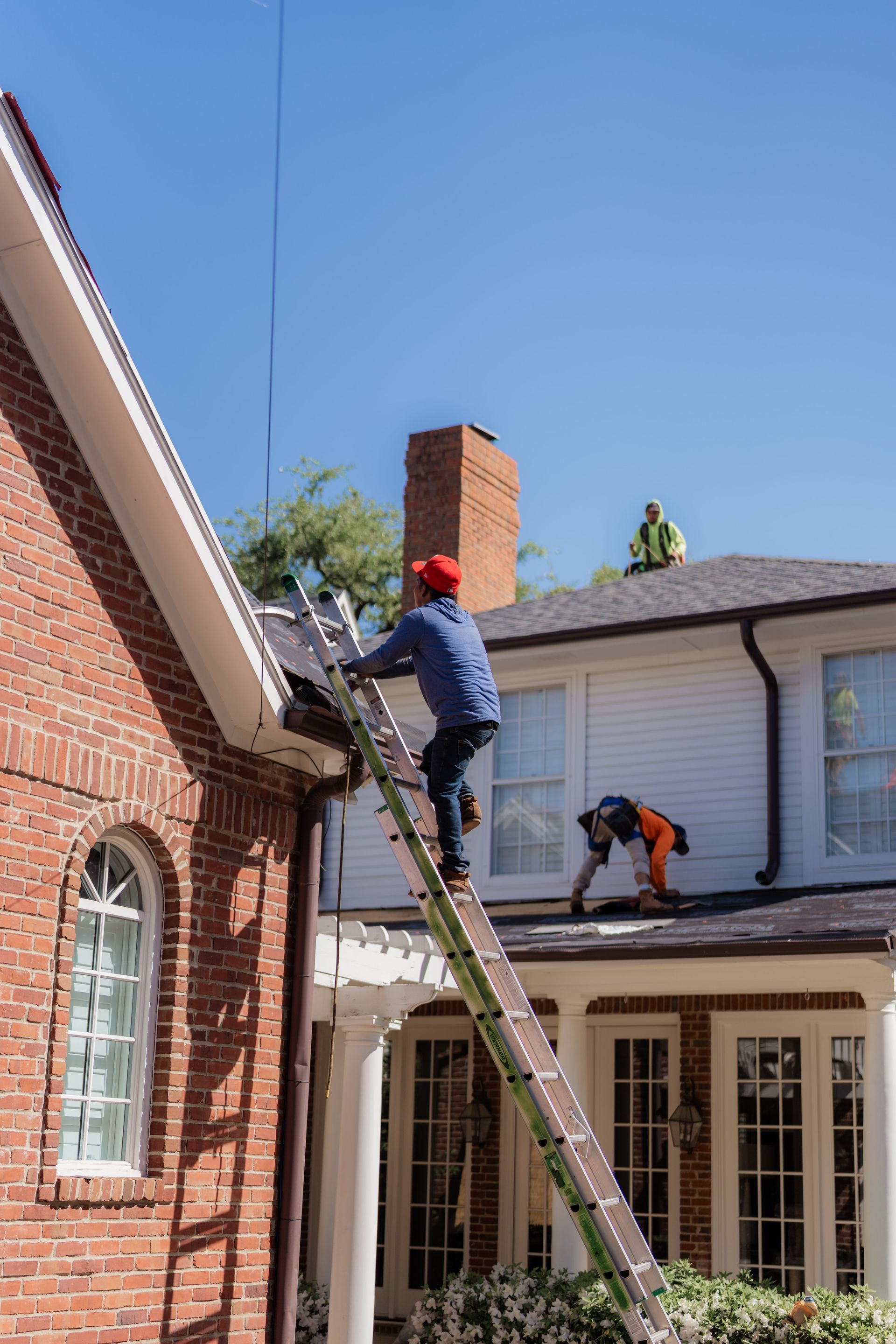 Construction workers on a roof and ladder, repairing a white house with a red brick chimney. Sunny day.