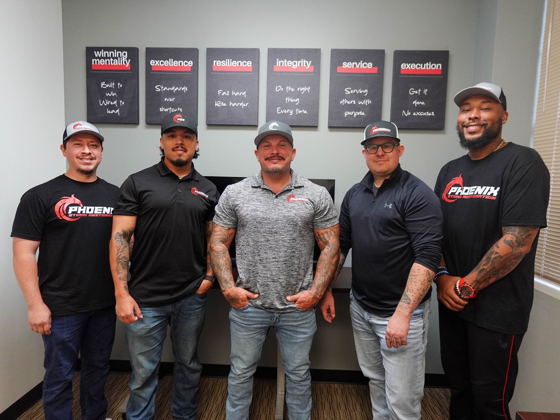 Five men in a room posing in front of wall decor. They all have hats and matching shirts with a company logo.