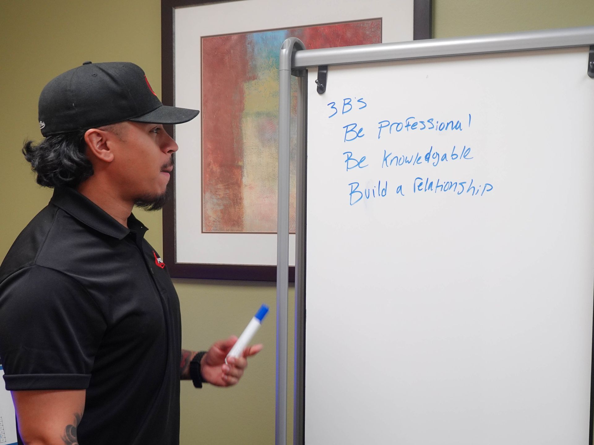 Man in black cap and shirt, points to whiteboard with