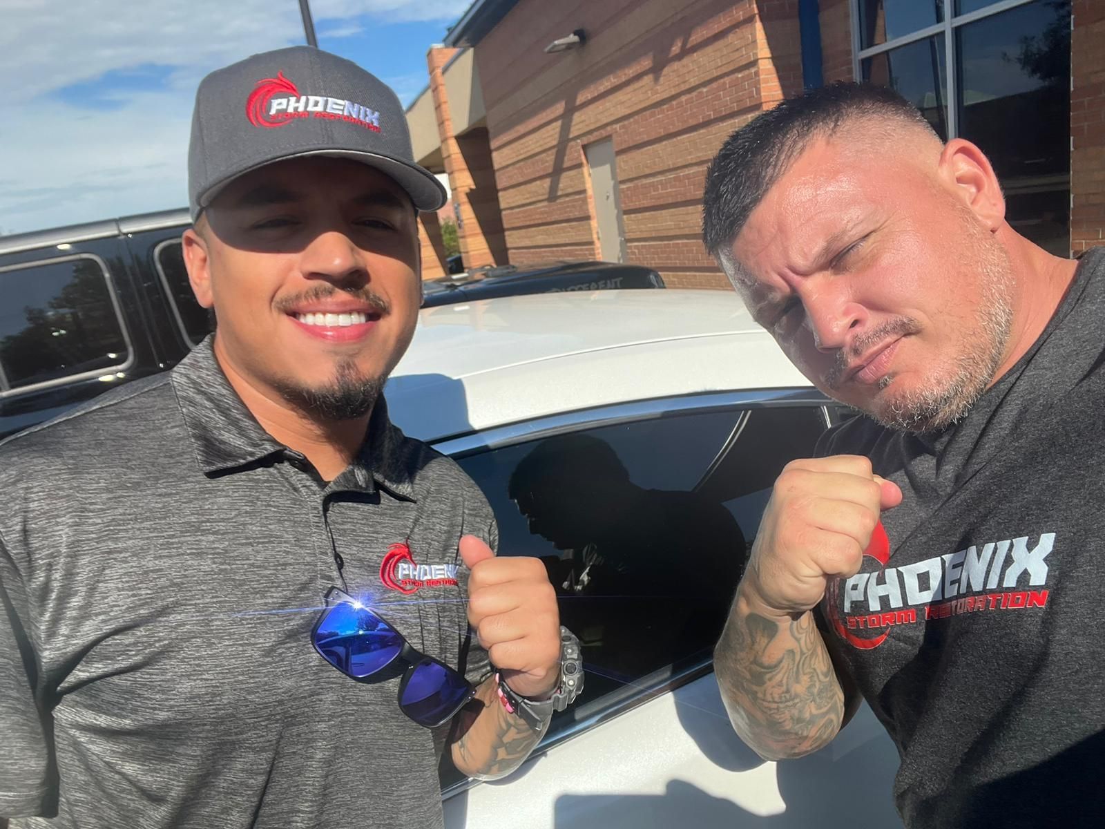 Two men smiling, giving thumbs-up, in grey Phoenix uniforms, by a car, sunny setting.