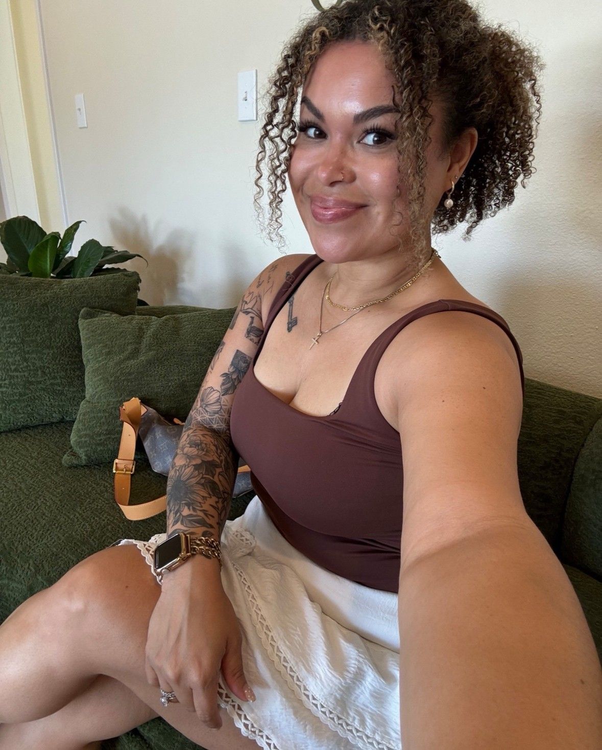 Woman with curly hair smiles, wearing a brown tank top and white skirt, seated on a green couch.