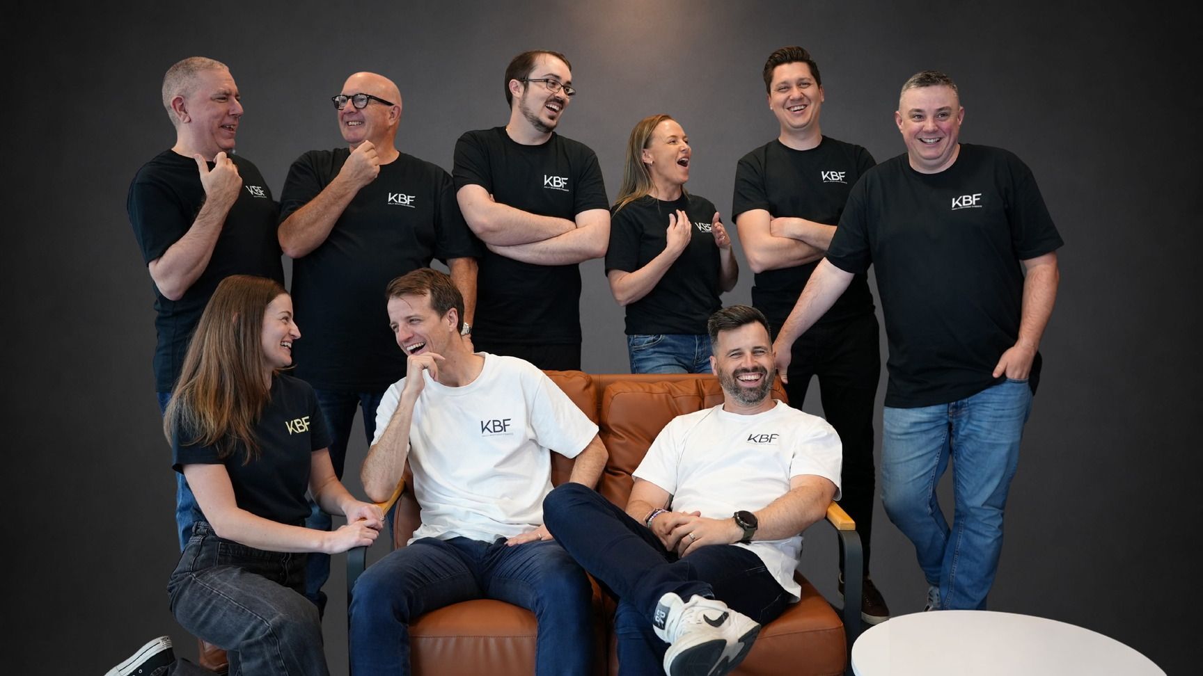 Group of people in black shirts posing and chatting on stools against a dark backdrop