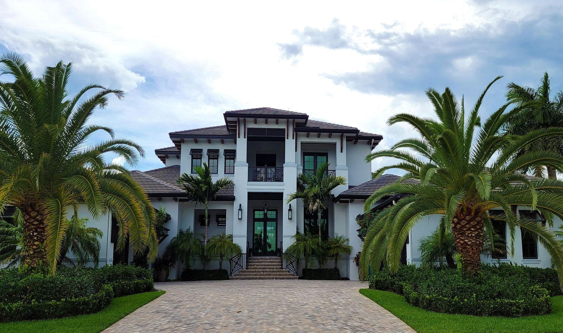 Large white tropical villa with palm trees and a stone driveway under a cloudy sky