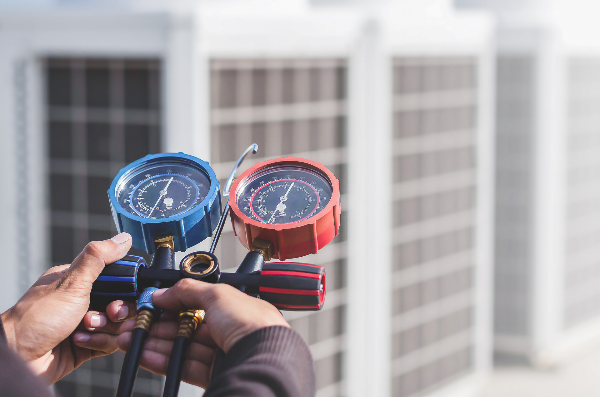 Hands holding blue and red HVAC gauges in front of rooftop air-conditioning units