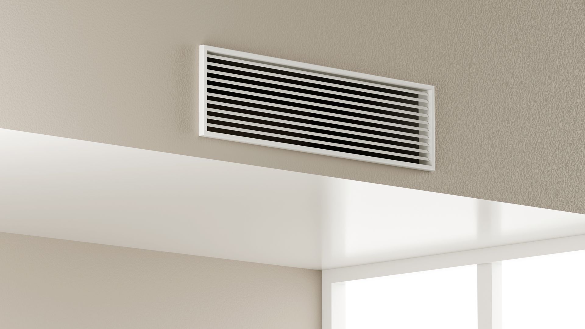 A rectangular white HVAC air vent mounted on a beige interior wall near a corner and a window.