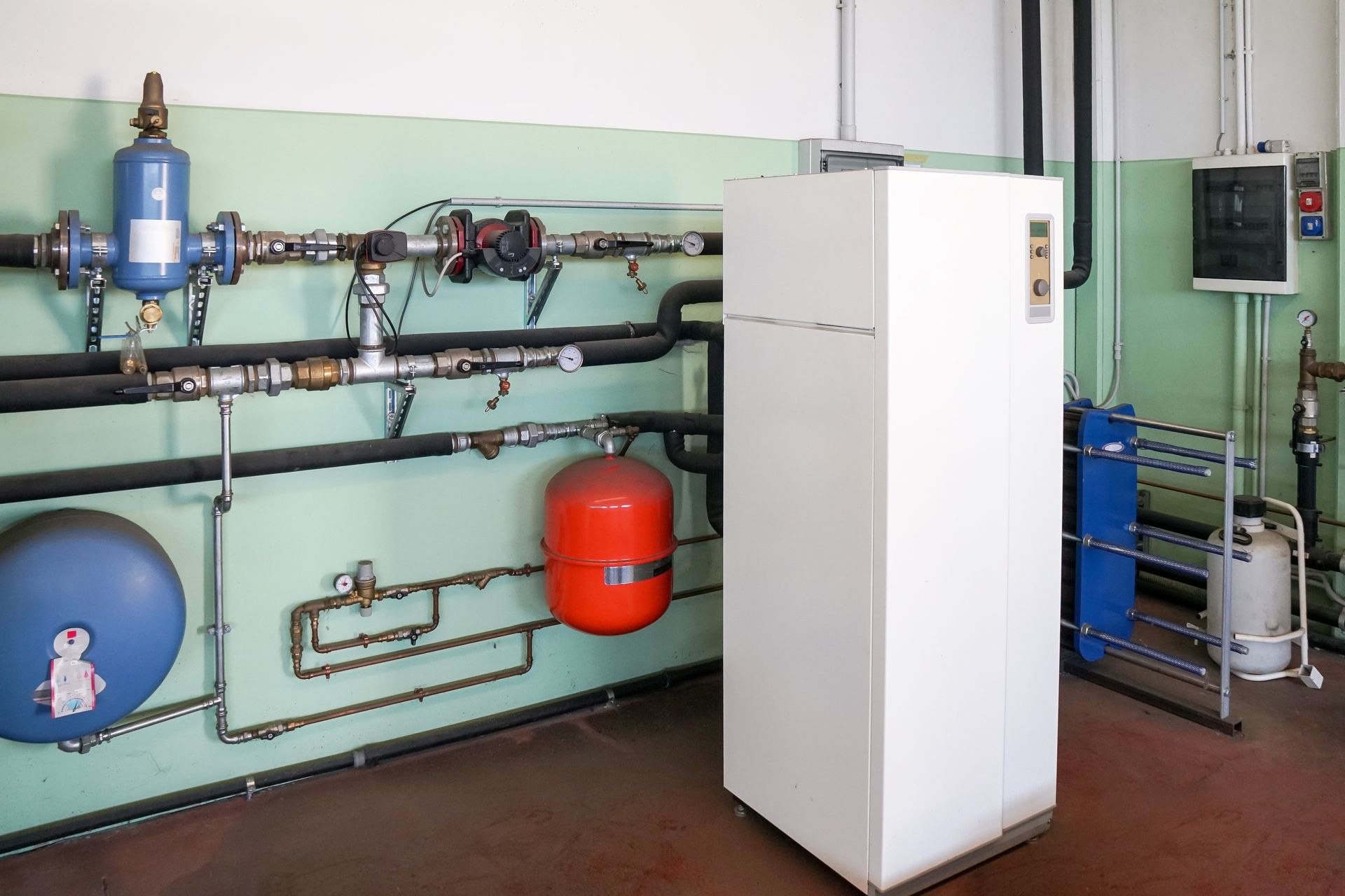 A mechanical room featuring a white heat pump, an orange expansion tank, a blue pressure vessel, and associated piping.