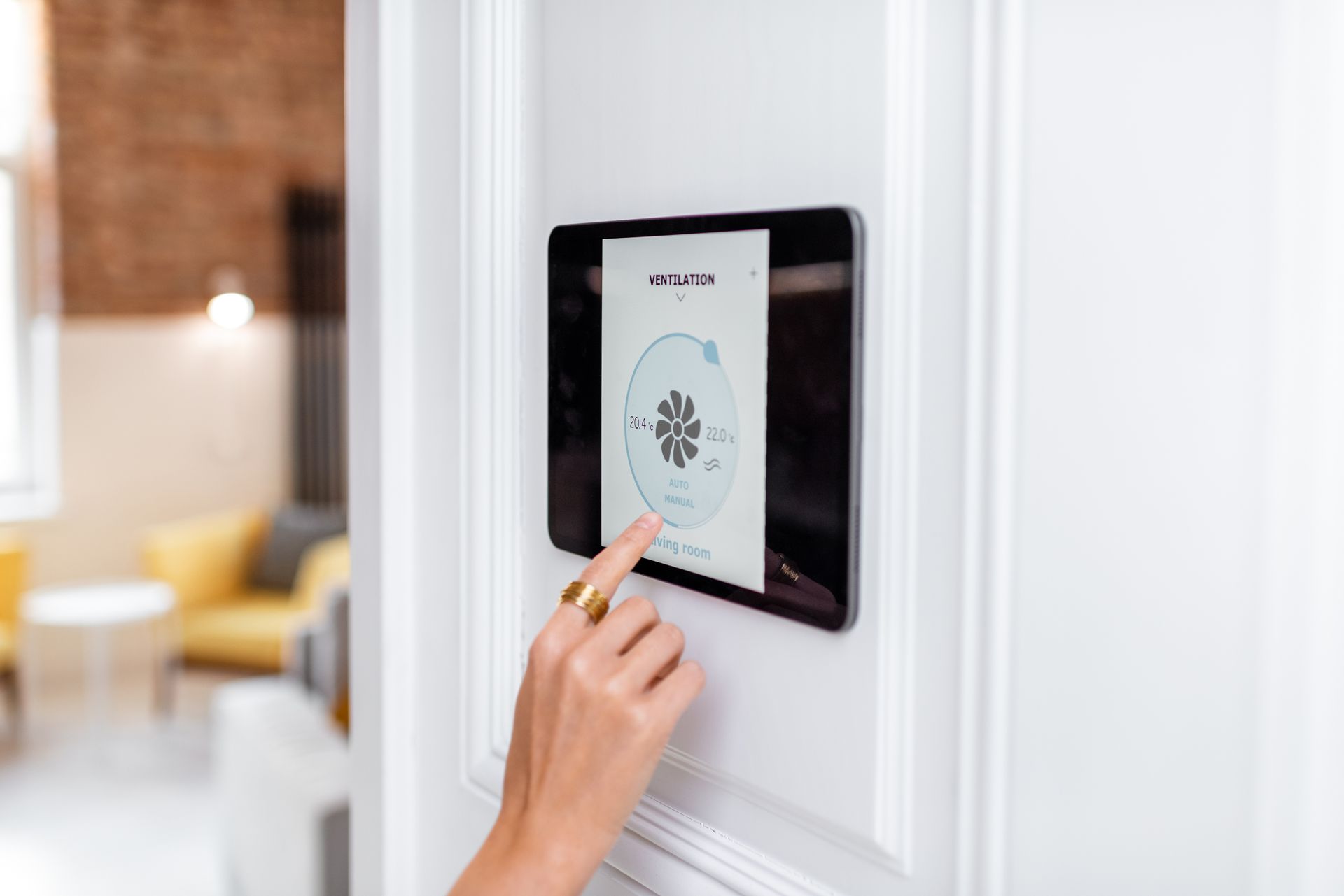 Person's hand touching a smart home control panel mounted on a white wall in a modern living space.