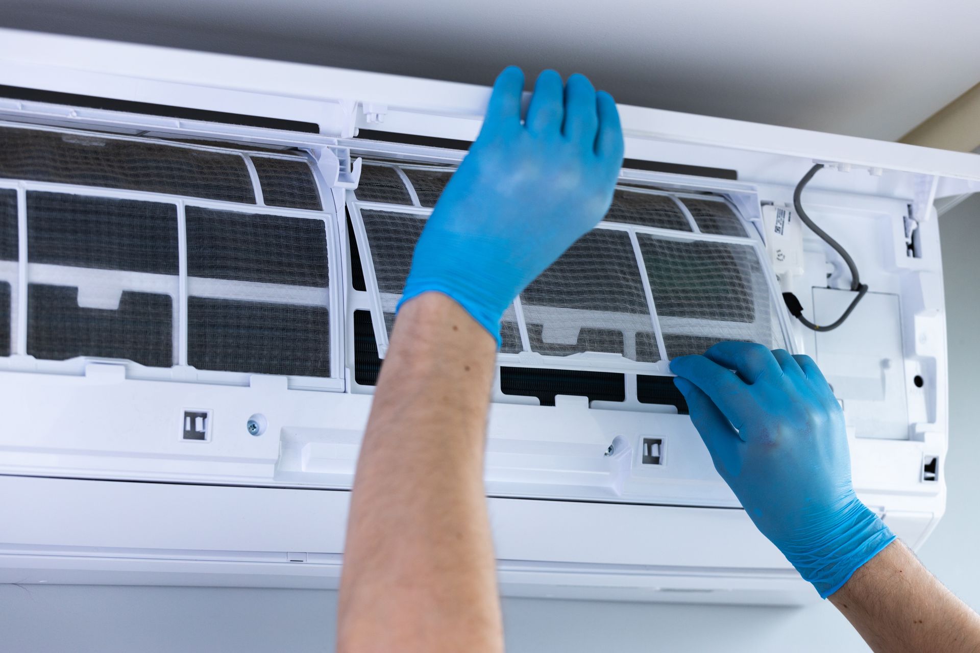 Hands in blue gloves cleaning the filter of a white air conditioner unit.