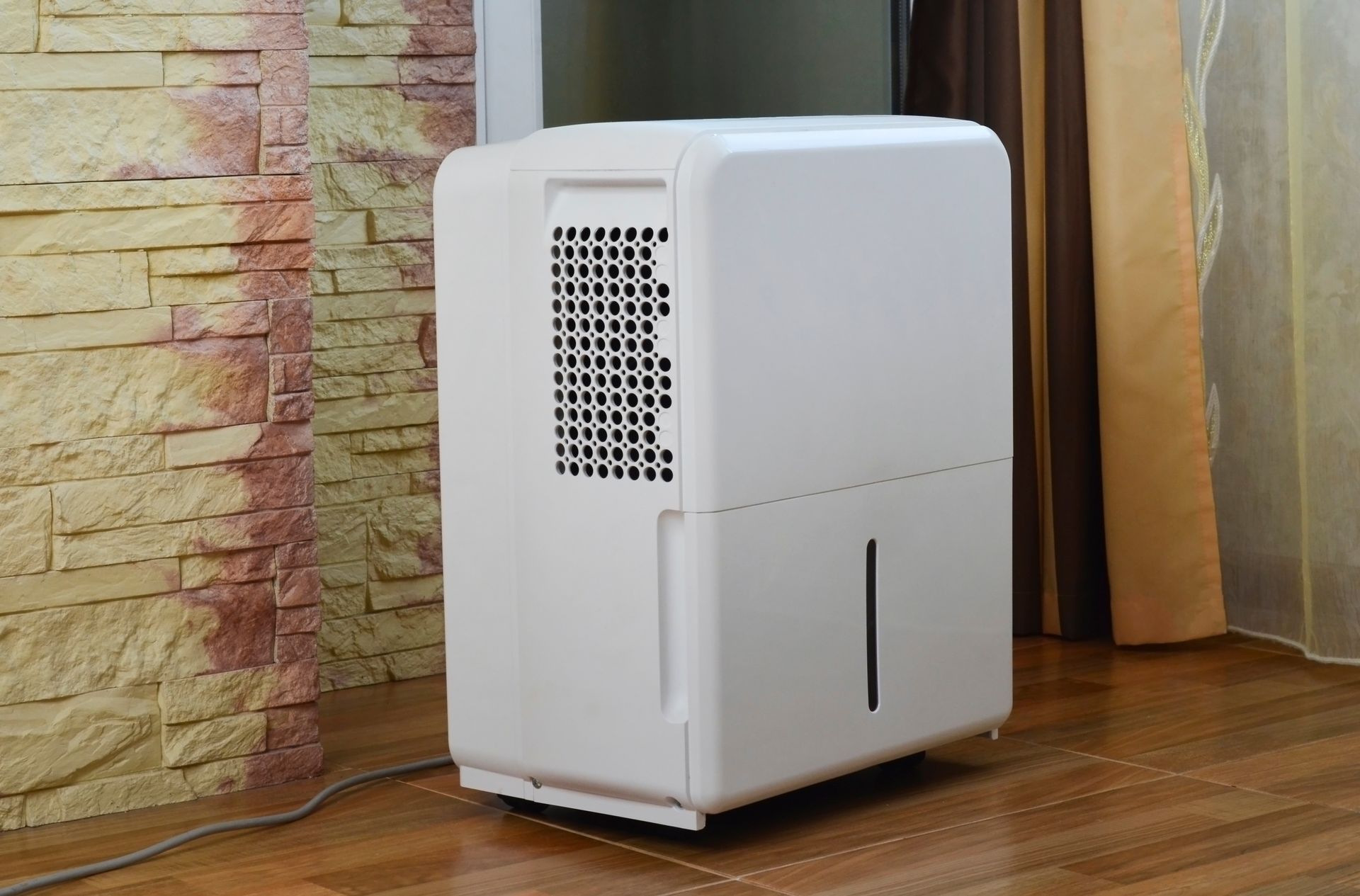 A white portable dehumidifier stands on a wooden floor in a room with a textured stone wall and curtains.