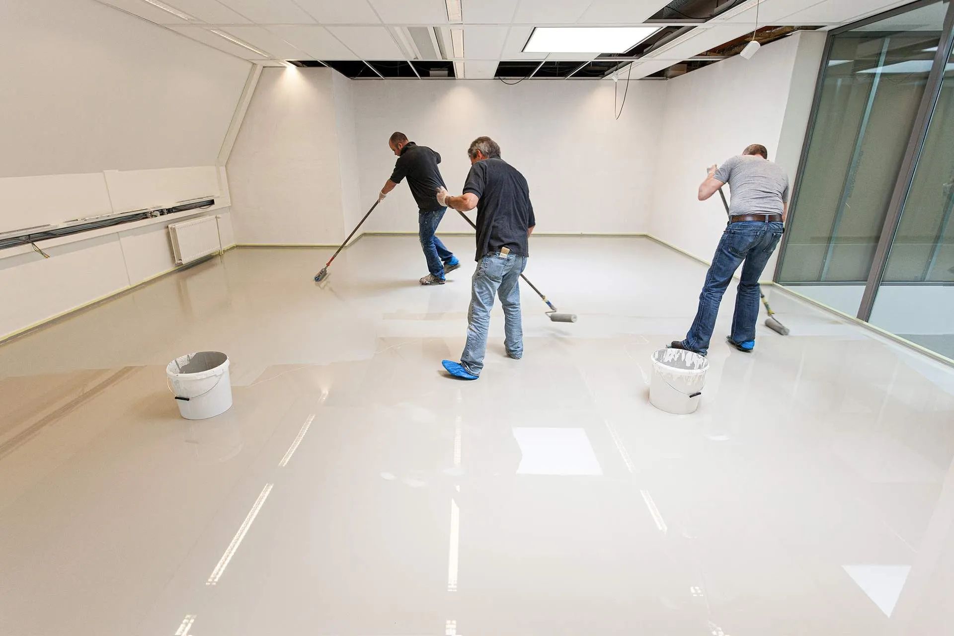 Workers applying glossy white coating to an empty room floor with rollers and paint buckets