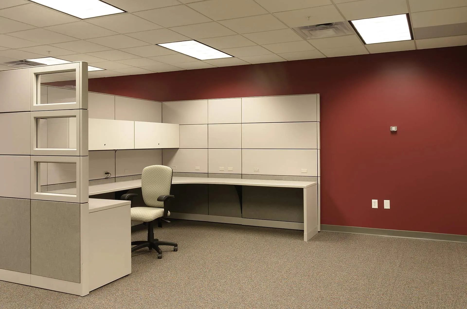 Empty office cubicle with white desks, beige chairs, maroon wall, and patterned carpet.