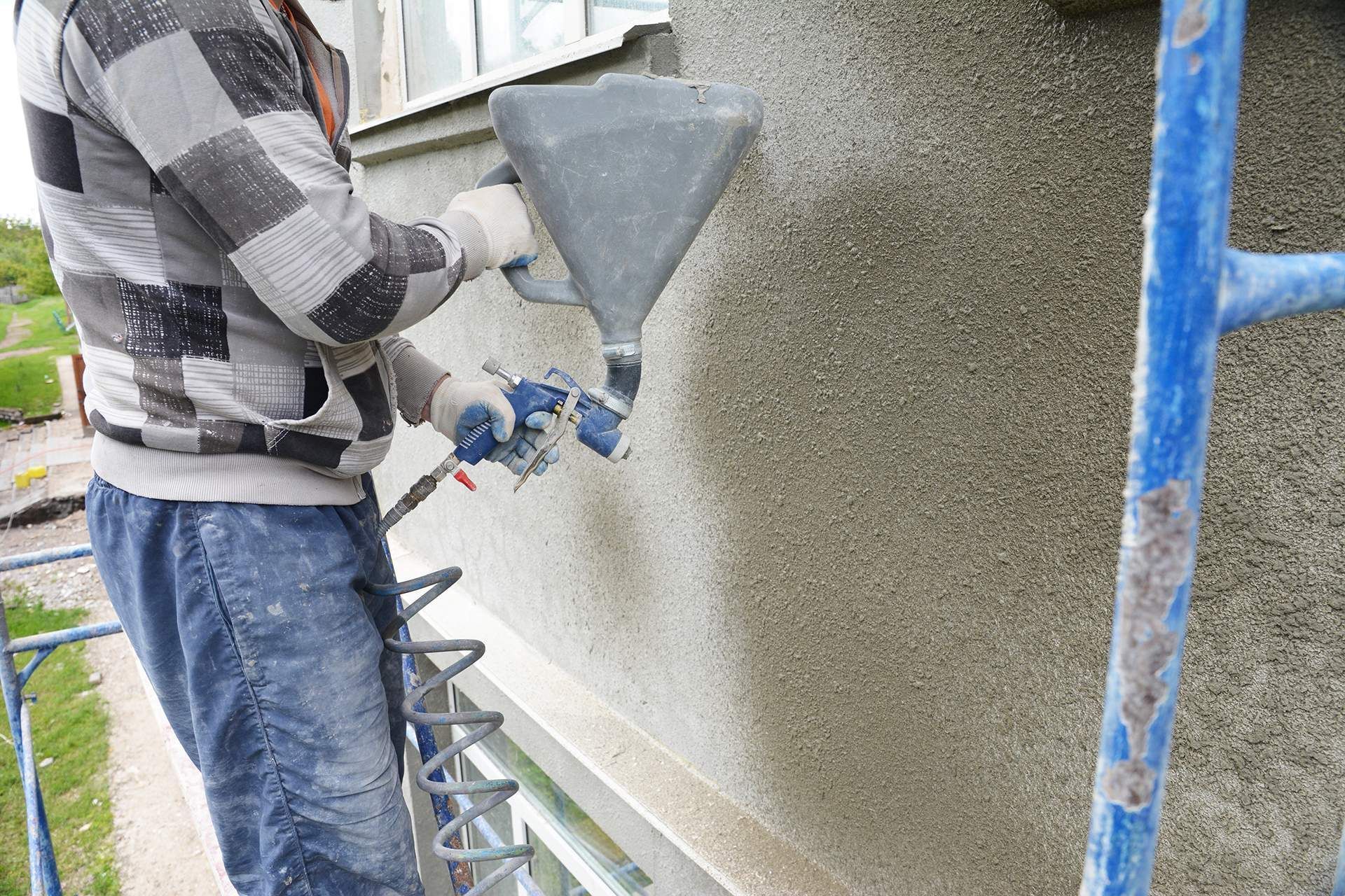 Worker spraying stucco onto an exterior wall beside blue scaffolding