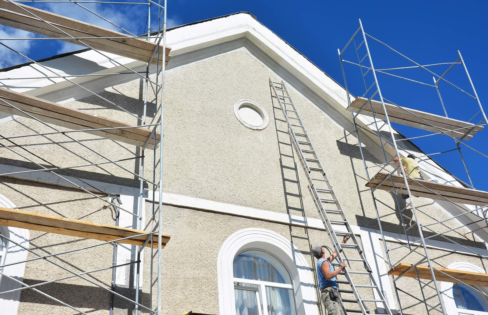 Scaffolding around a beige house as a worker climbs a ladder near a window