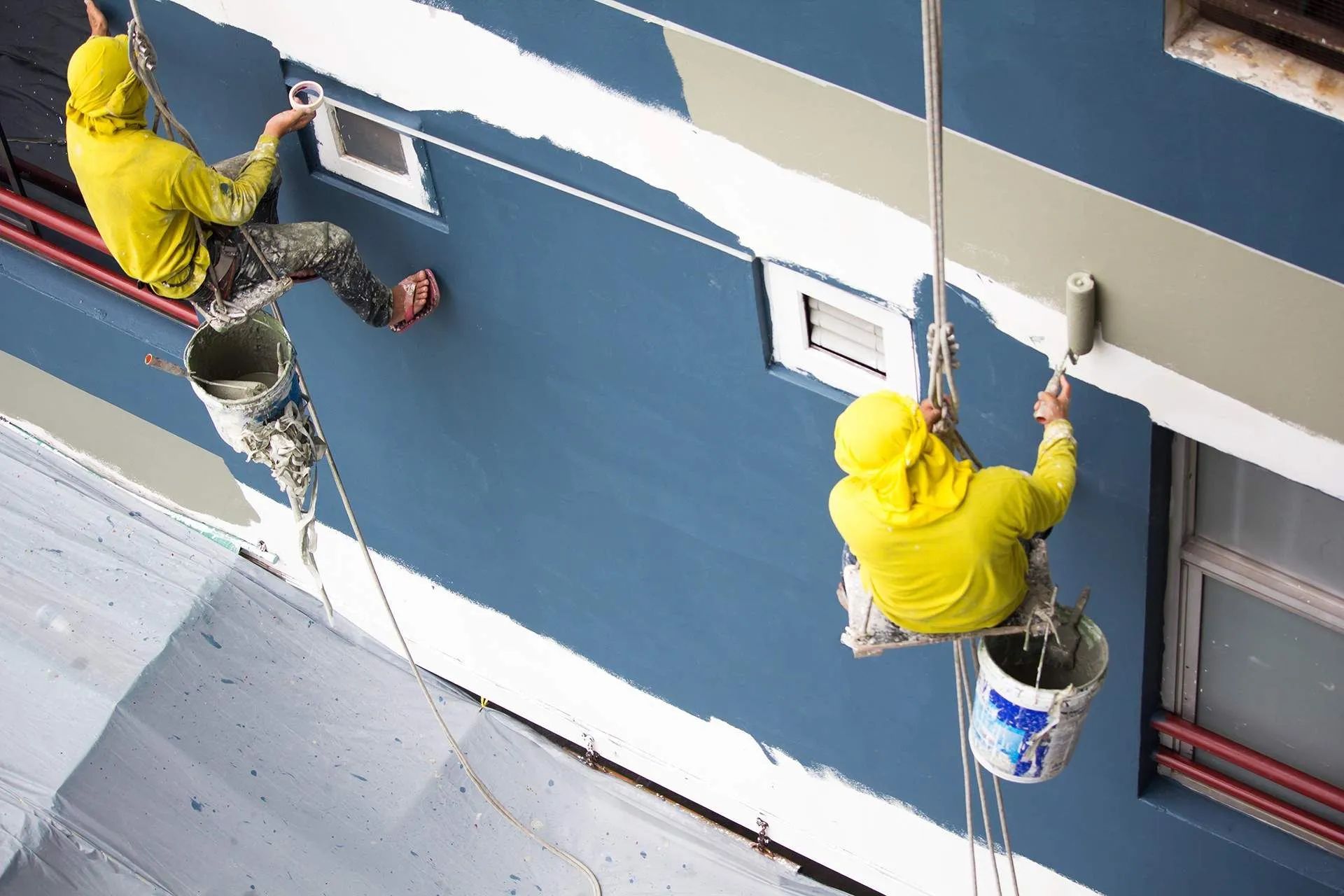 Two workers in yellow hoodies hang on ropes, painting a blue building wall with white trim.