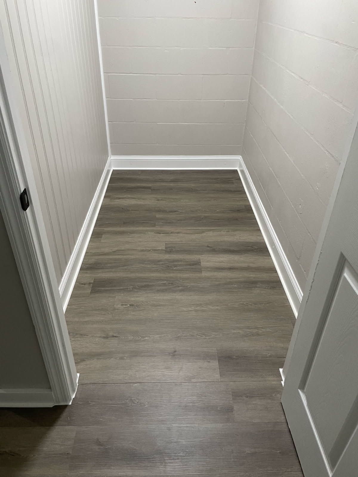 Narrow empty hallway with white walls and gray wood flooring, viewed through an open doorway