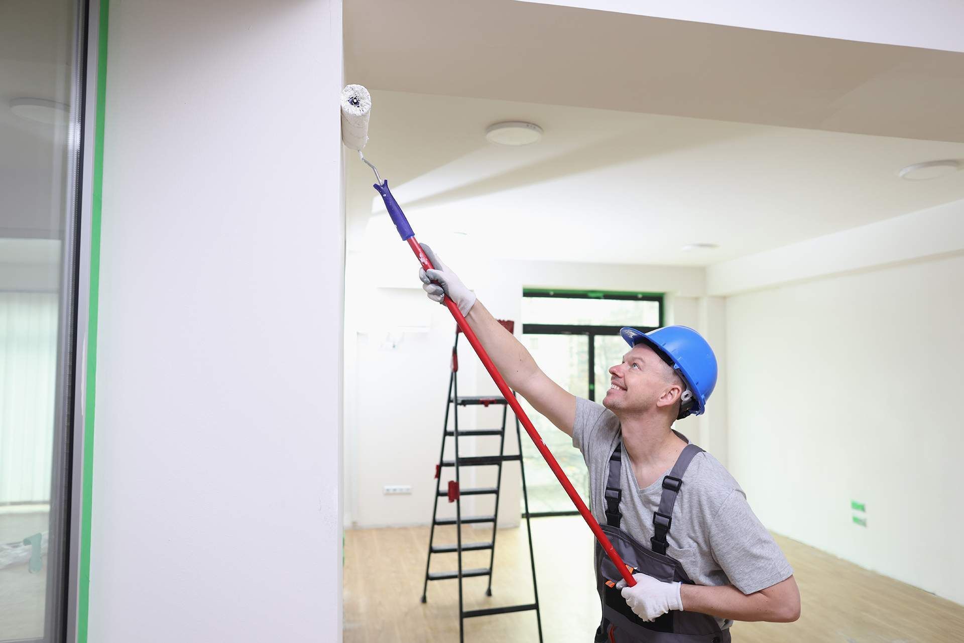 Worker in blue hard hat painting a ceiling with a long roller in a bright room