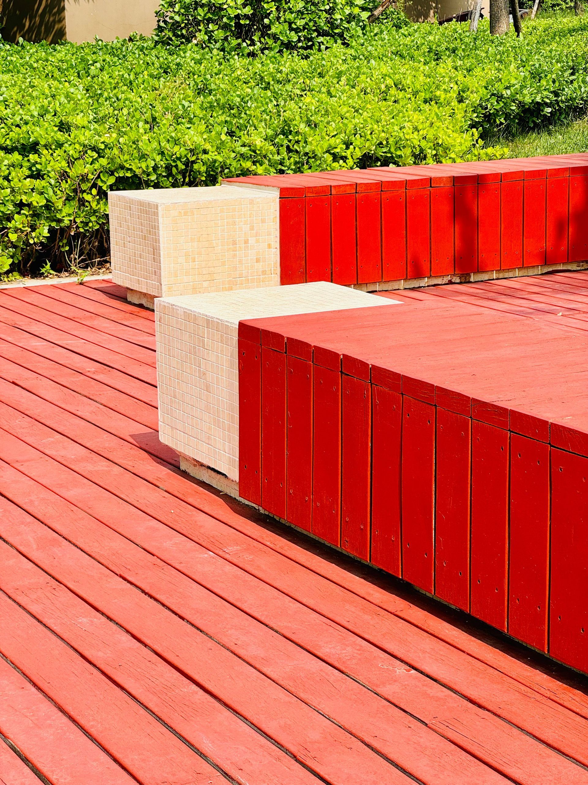 Red wooden benches on a sunny outdoor deck beside green hedges and beige stone blocks