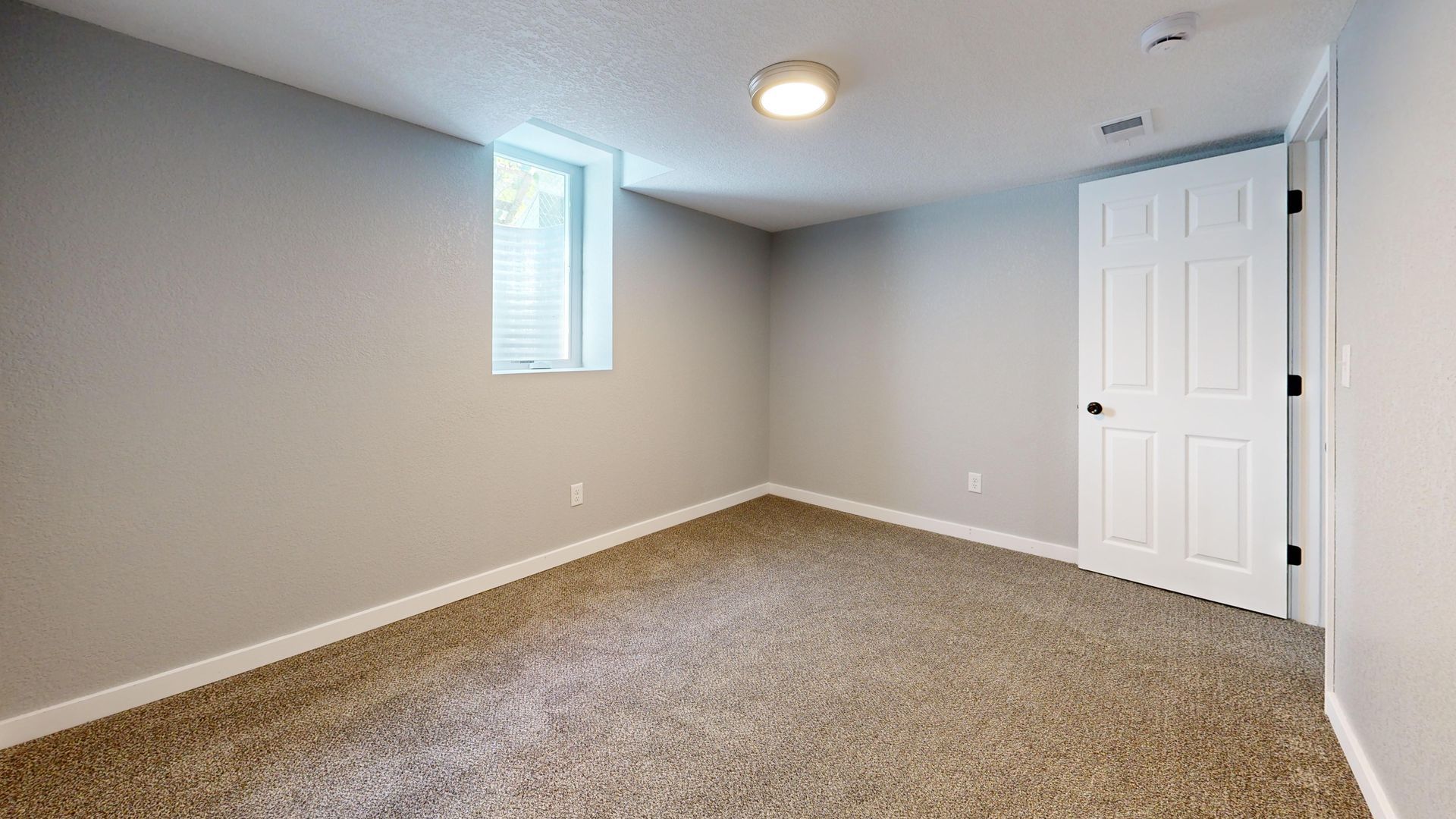 Empty beige carpeted room with gray walls, a small window, ceiling light, and white closet doors.