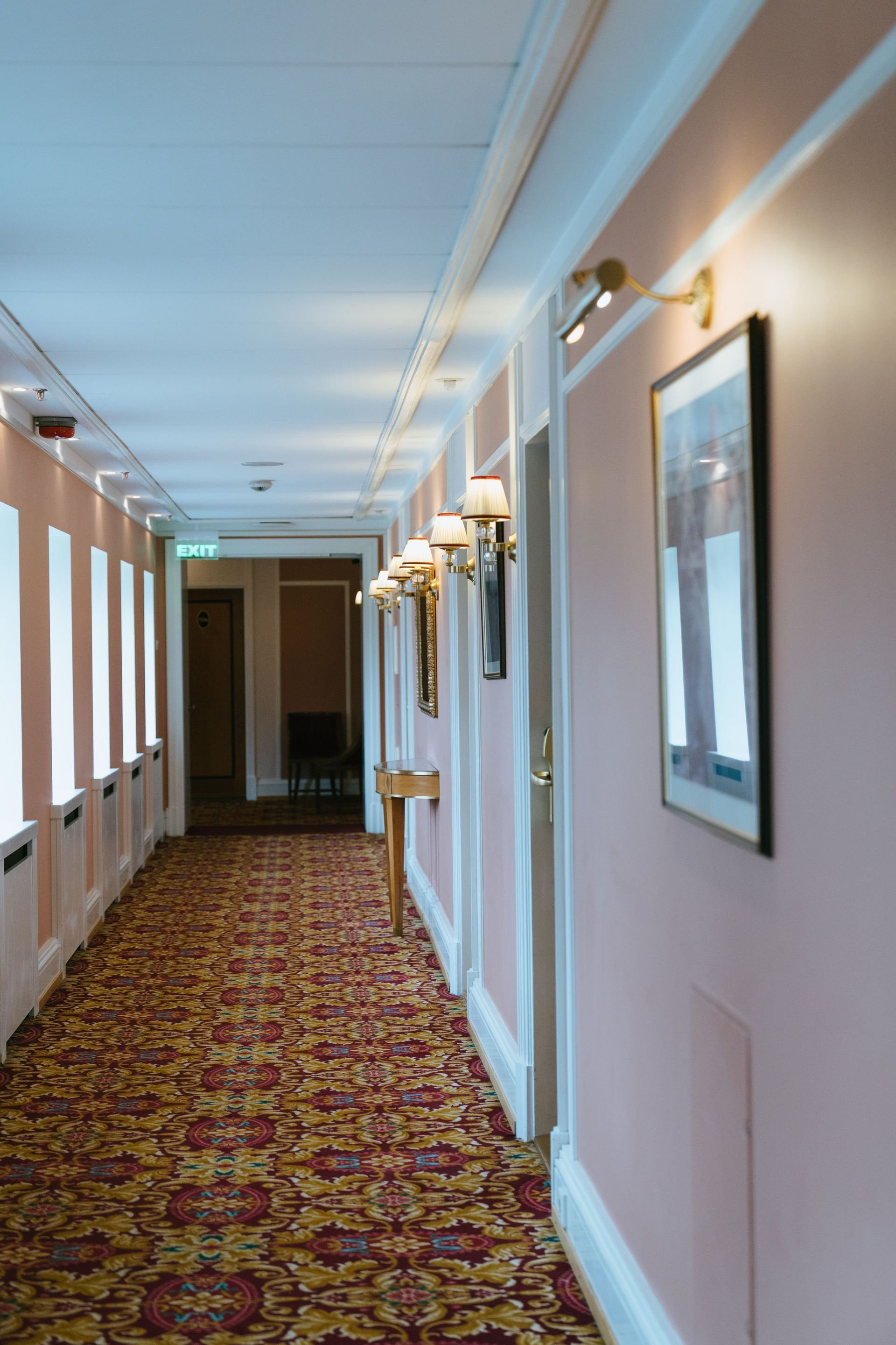 Hotel hallway with patterned carpet, wall sconces, and framed artwork along a pink corridor