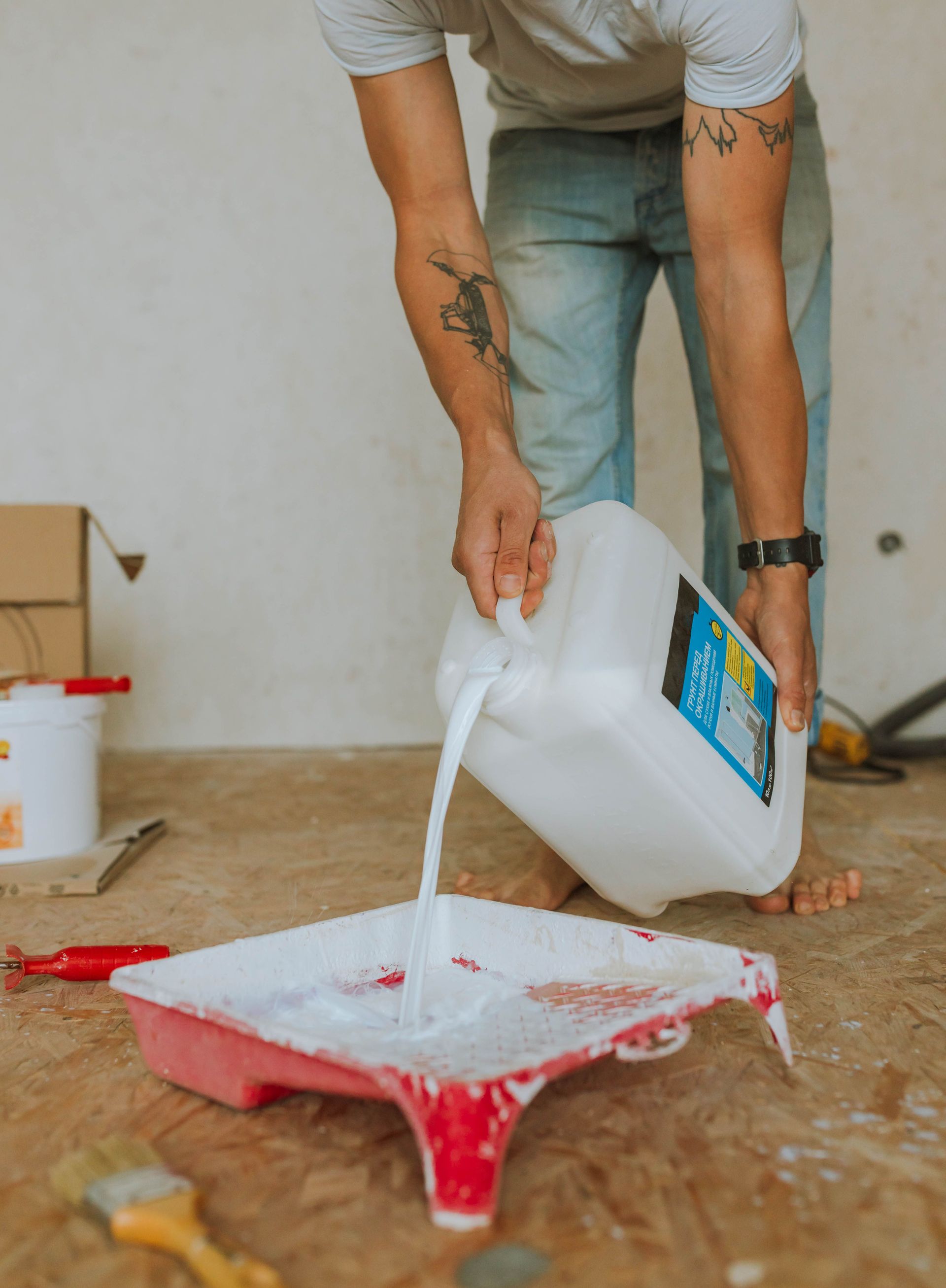 Person pours white paint from a jug into a red paint tray on a wooden floor.