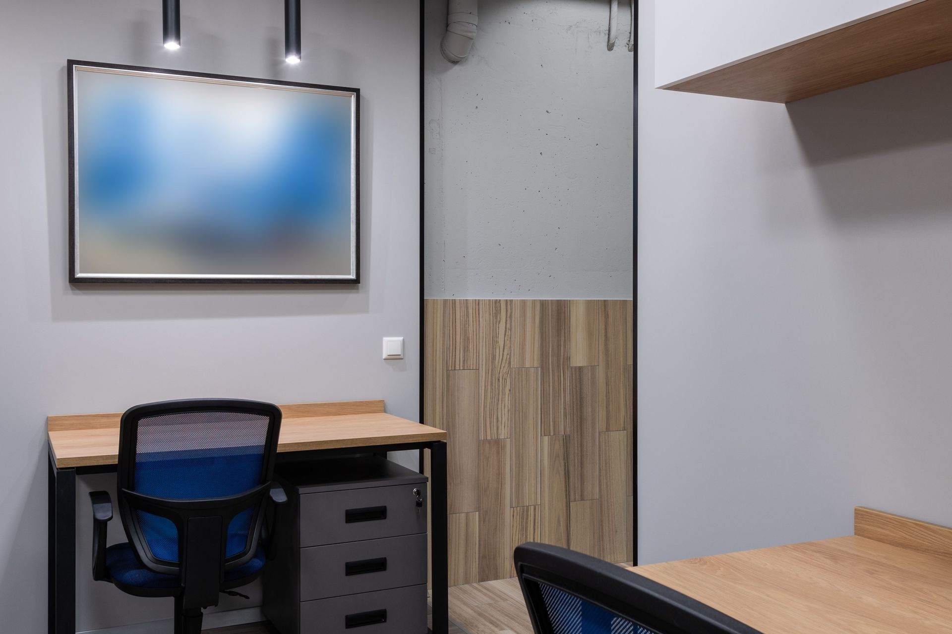 Empty office cubicle with wooden desks, blue chairs, and a framed abstract print on a gray wall