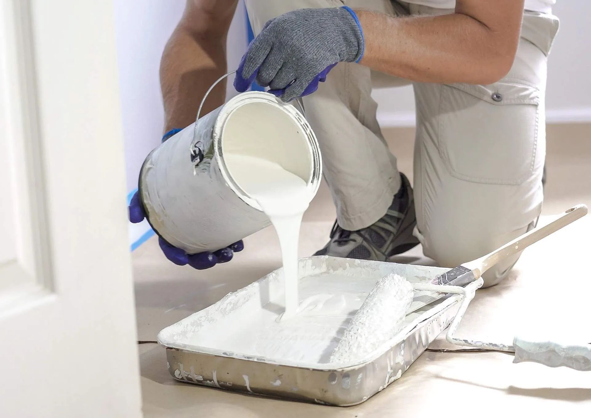 Person pours white paint into a tray while kneeling on a floor under renovation.