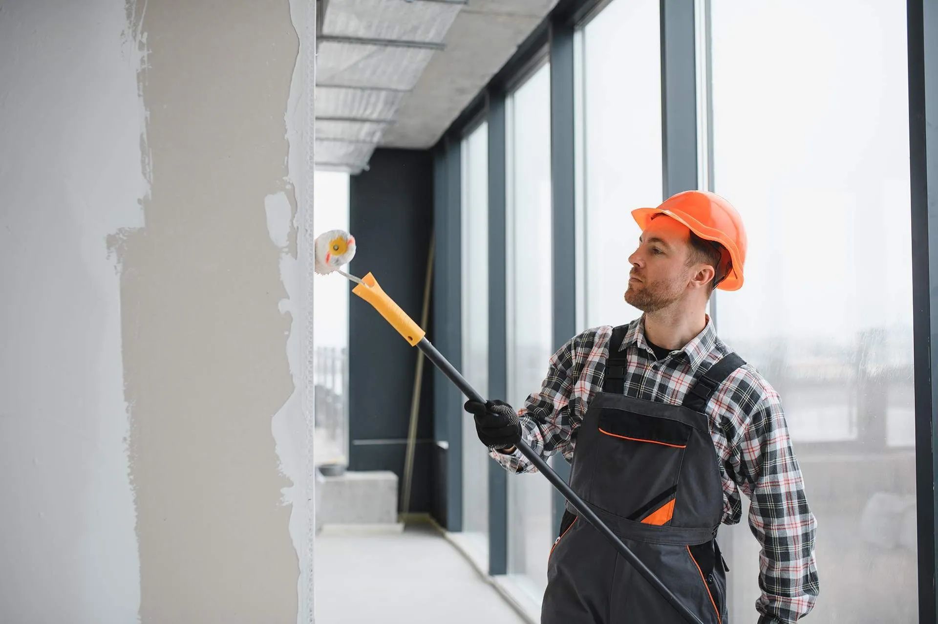 Worker painting an indoor wall with a roller in a bright hallway, wearing orange safety gear and overalls