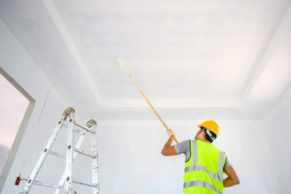 Worker in a yellow vest and hard hat painting a white ceiling beside a ladder