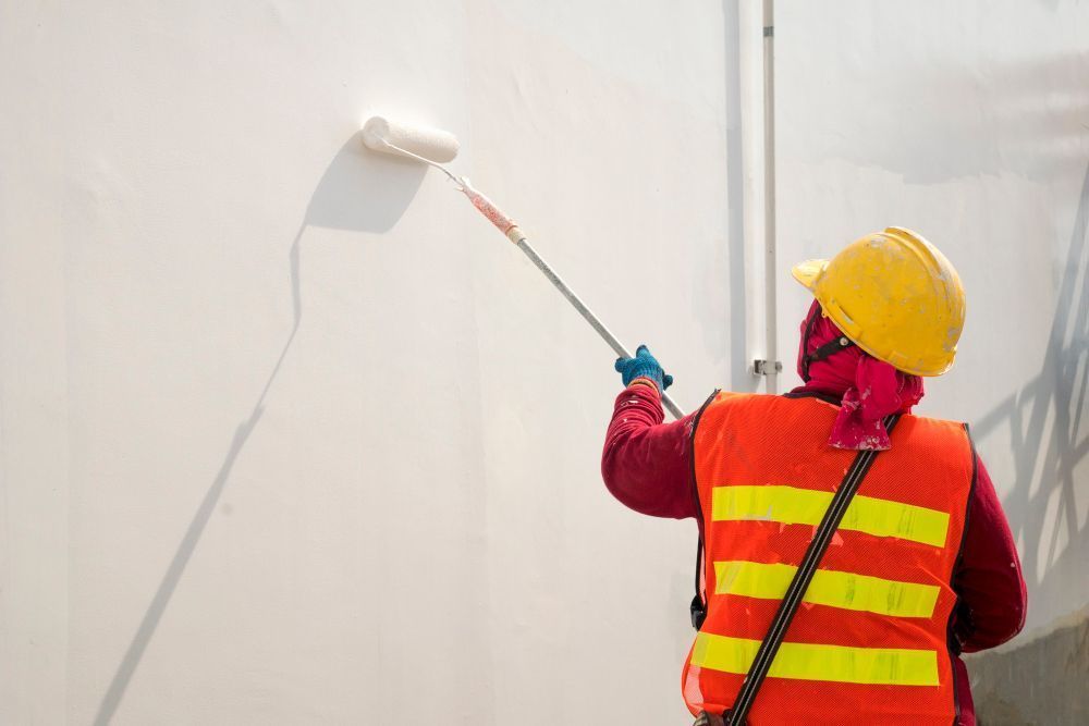 Worker in yellow hard hat and orange vest painting a wall with a roller