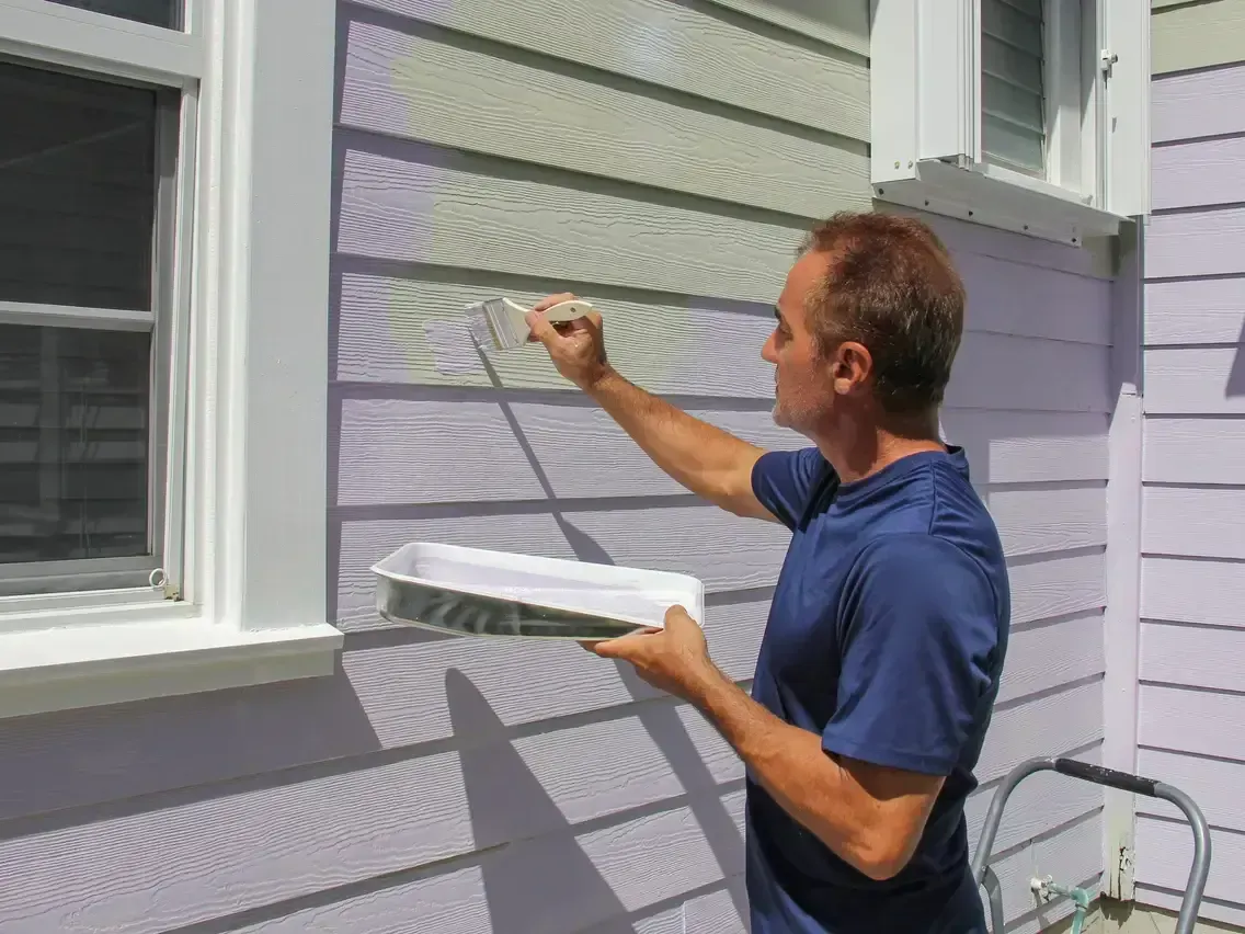Man painting a gray house exterior while holding a paint tray
