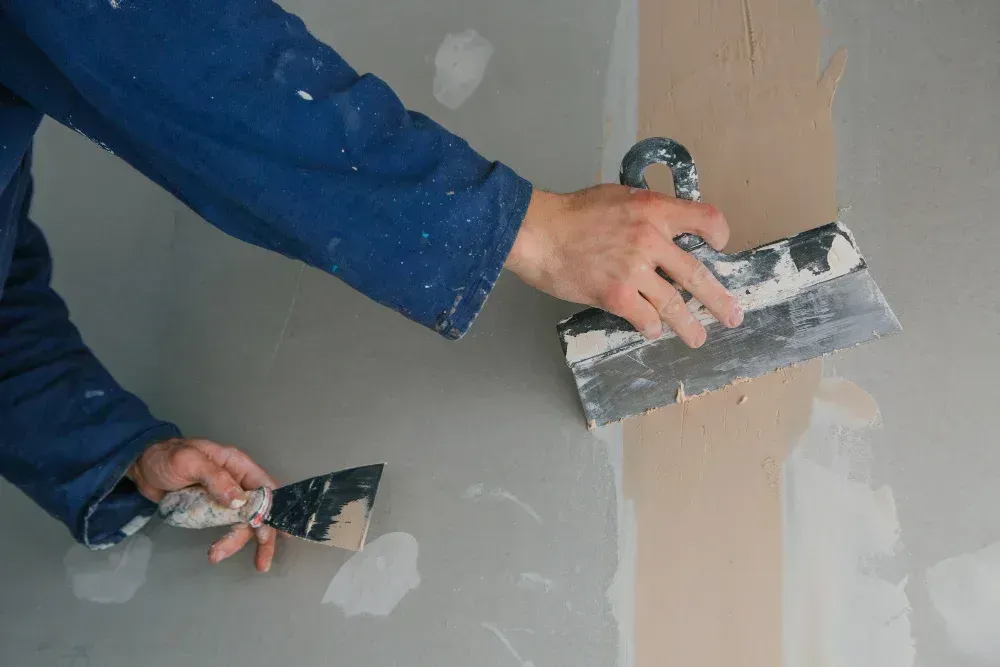 Hands using putty knives to apply drywall compound on a wall patch.
